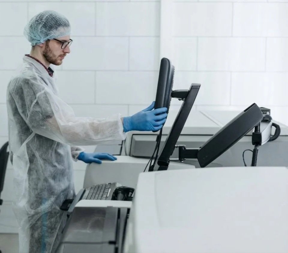 A scientist in protective gear, including a hair cover, gloves, and a lab coat, operating a computer in a clean laboratory environment.