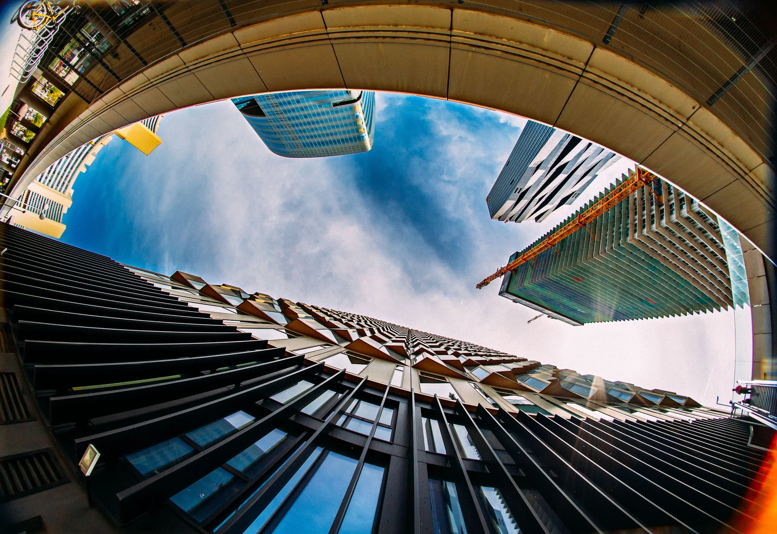 fisheye view of multiple skyscrapers, with a curved pedestrian bridge overhead, in an urban cityscape. The sky is partly cloudy.