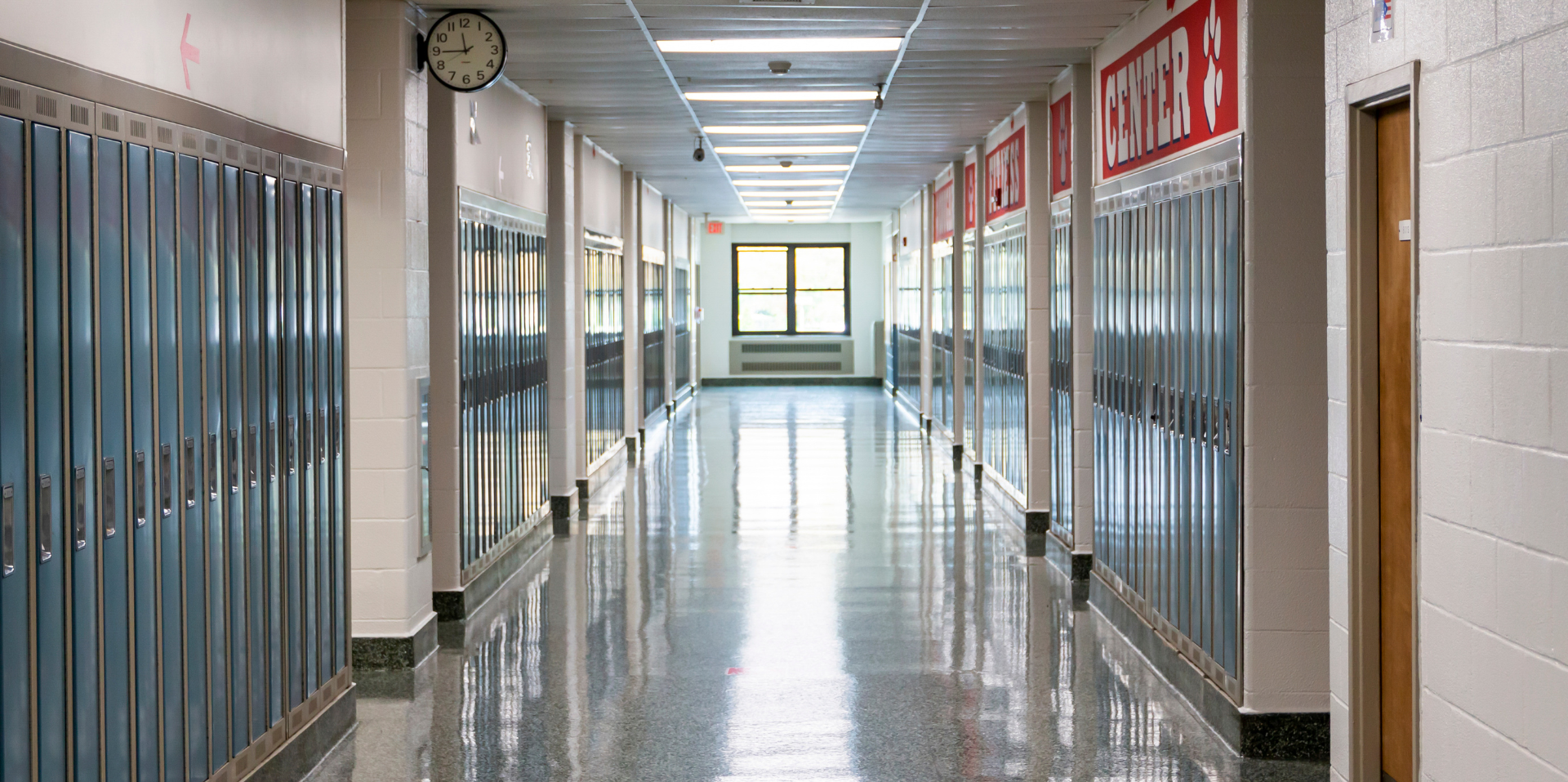 Empty school hallway with blue lockers on both sides, a clock on the wall, and a window at the end, brightly lit with overhead lights.
