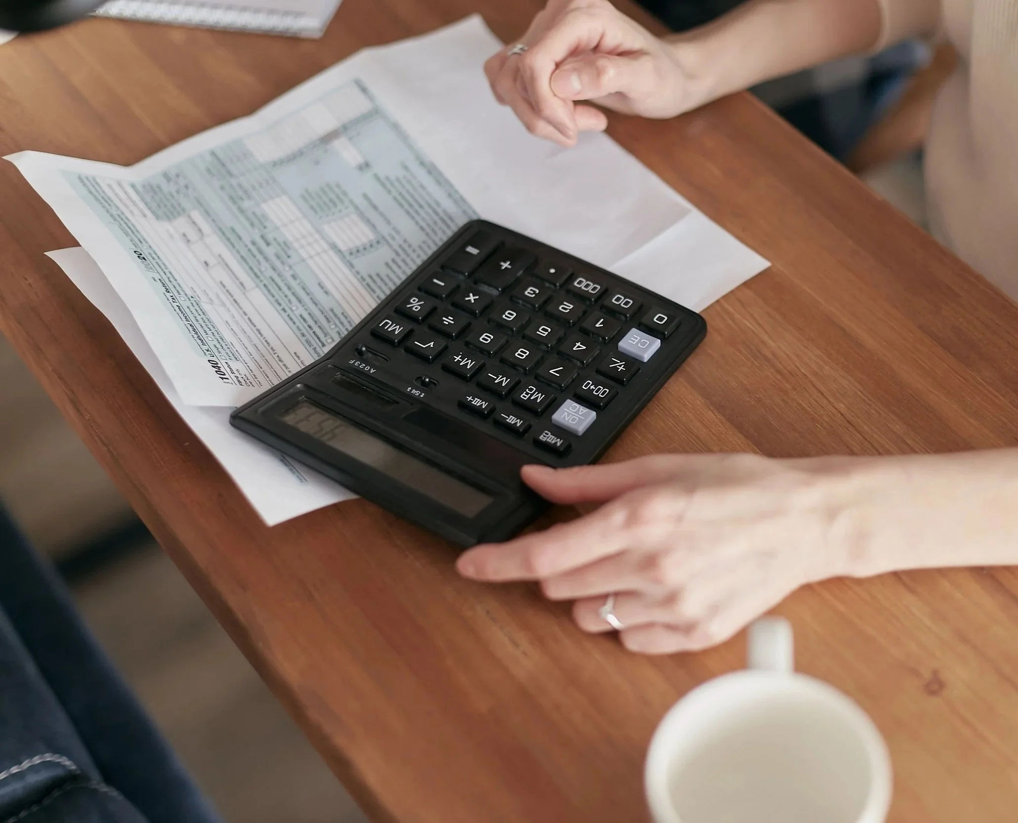 Hands resting on a wooden table with paper and a calculator, possibly doing financial work or calculations.
