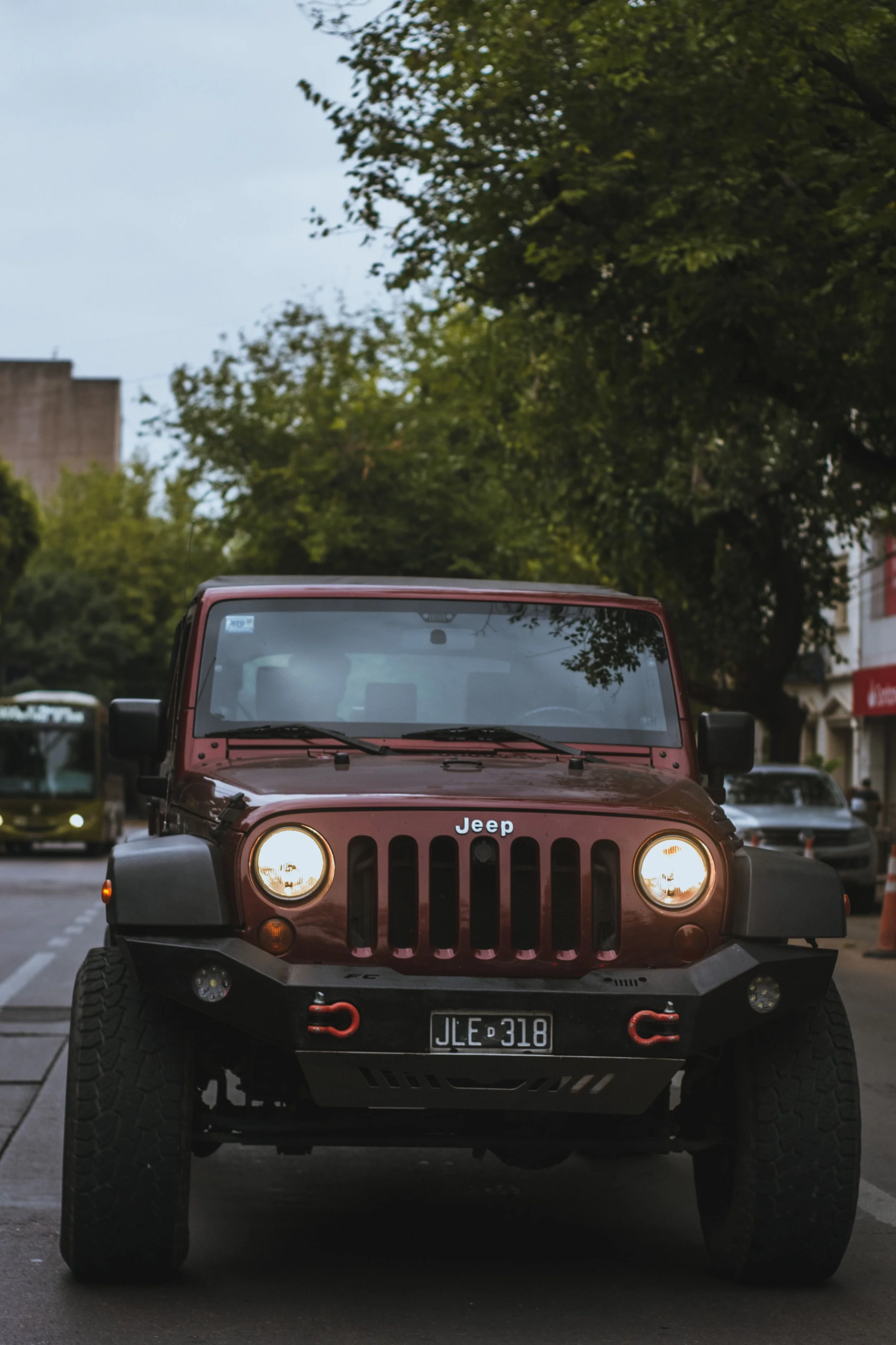 Front view of a red Jeep Wrangler with its headlights on, parked on a city street lined with trees and buildings.