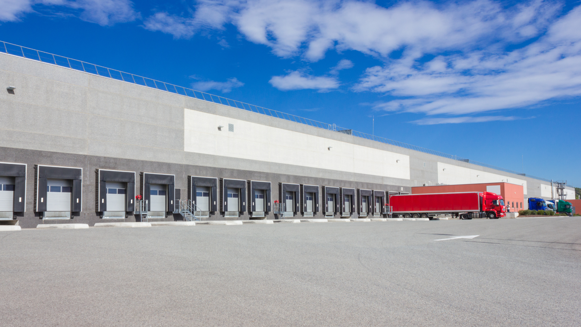 A large warehouse with multiple loading docks, three trucks parked in front, and a bright blue sky with some clouds.