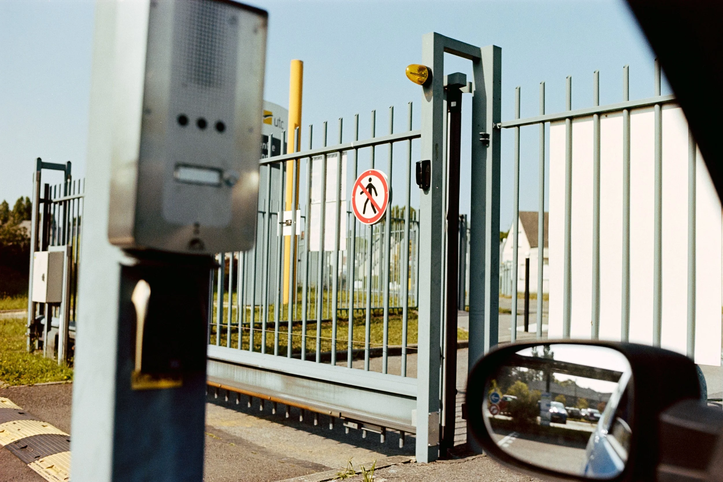 A metal gate with a security device in front, viewed from a car with a side mirror reflecting the scene. The gate has a sign indicating no pedestrians are allowed, and a yellow warning light is attached near the top.