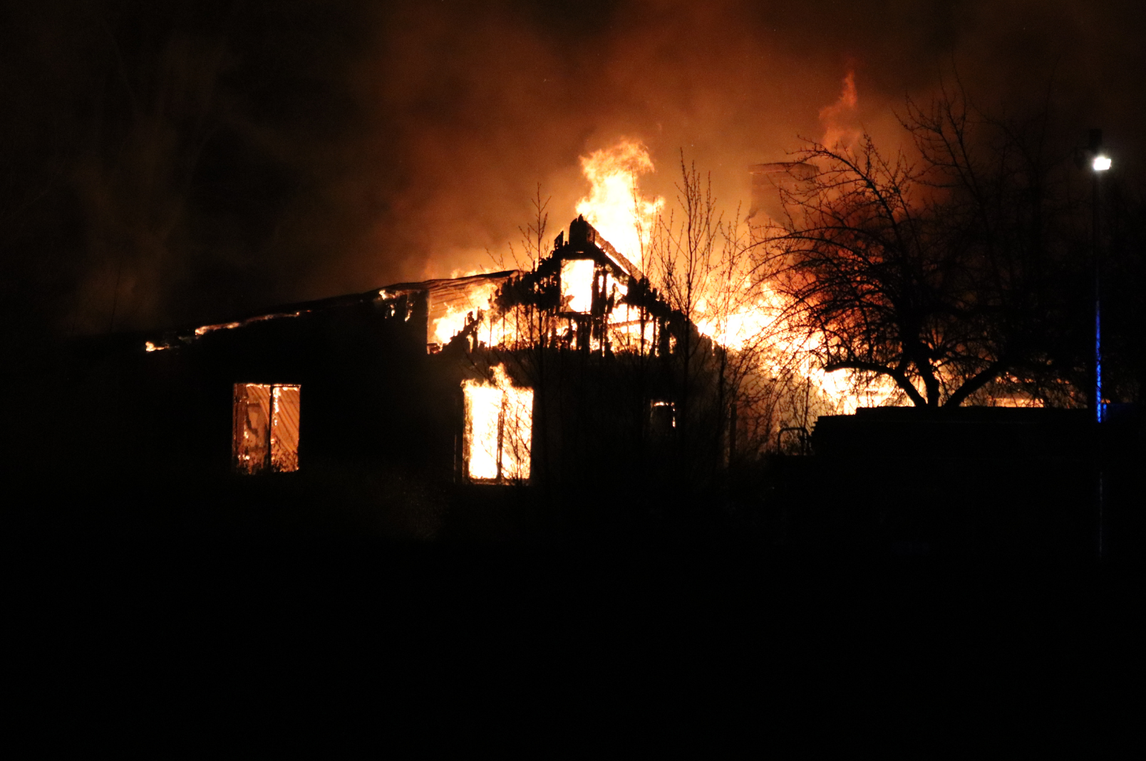 A house engulfed in flames during a fire at night, with bright orange and yellow flames illuminating the dark surroundings and leafless trees in the foreground.