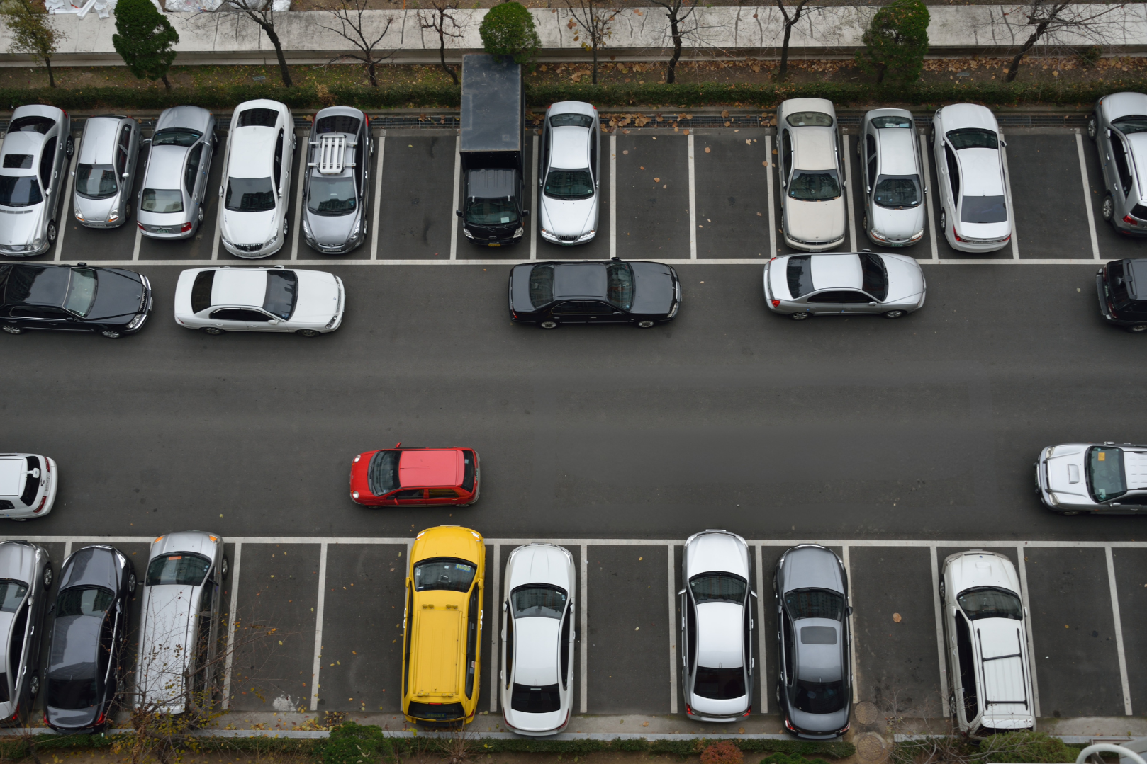 An aerial view of a parking lot with rows of parked cars and a few empty spaces, a narrow street running parallel with moving cars, and trees along the sidewalk.