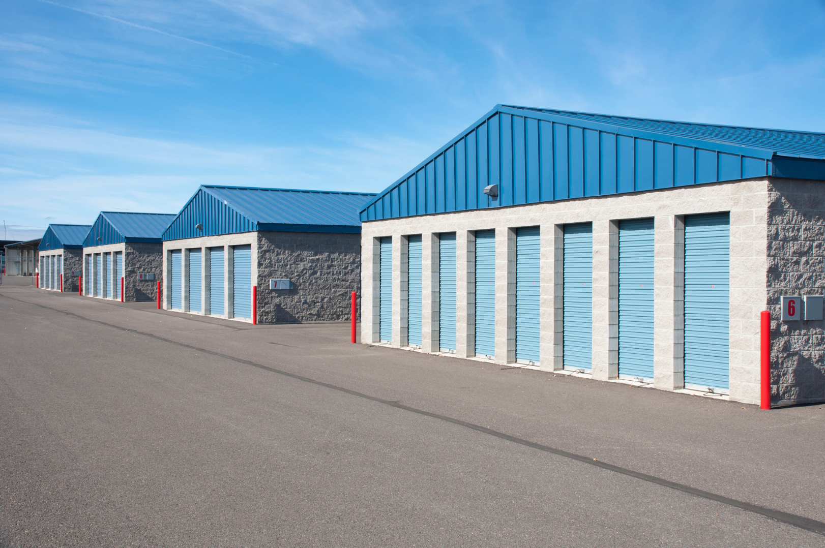 Row of storage units with blue roll-up doors and blue metal roofs on a paved lot, under a clear blue sky.