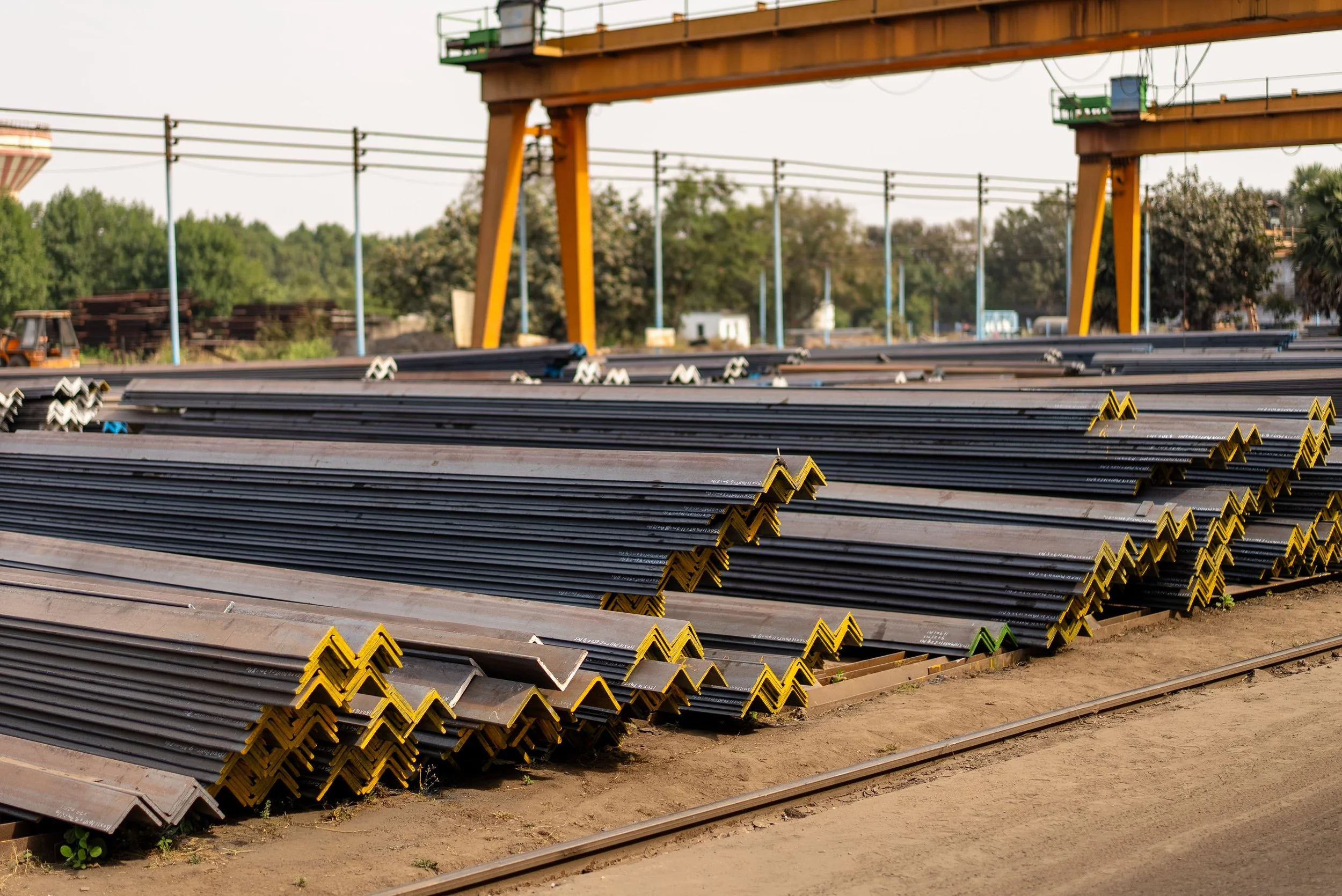 Stacks of steel rails with yellow ends stacked on the ground in an industrial area, with construction equipment, trees, and utility poles in the background.