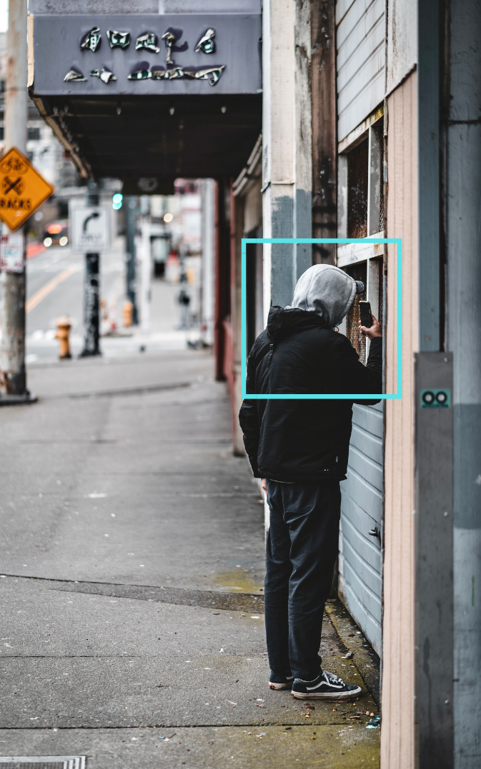 A person wearing a hoodie and black jacket standing on a city sidewalk, looking into a business window with a blue box outlined indicating suspicious activity detected through ai security feed.