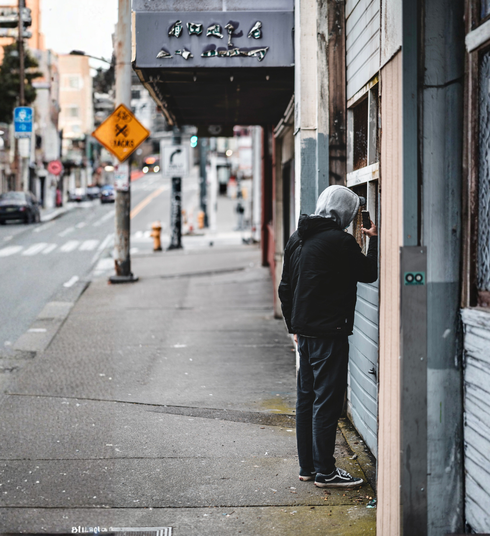 A person wearing a gray hoodie and black jacket takes a photo through a window on an urban sidewalk, with city streets, buildings, and signs in the background.