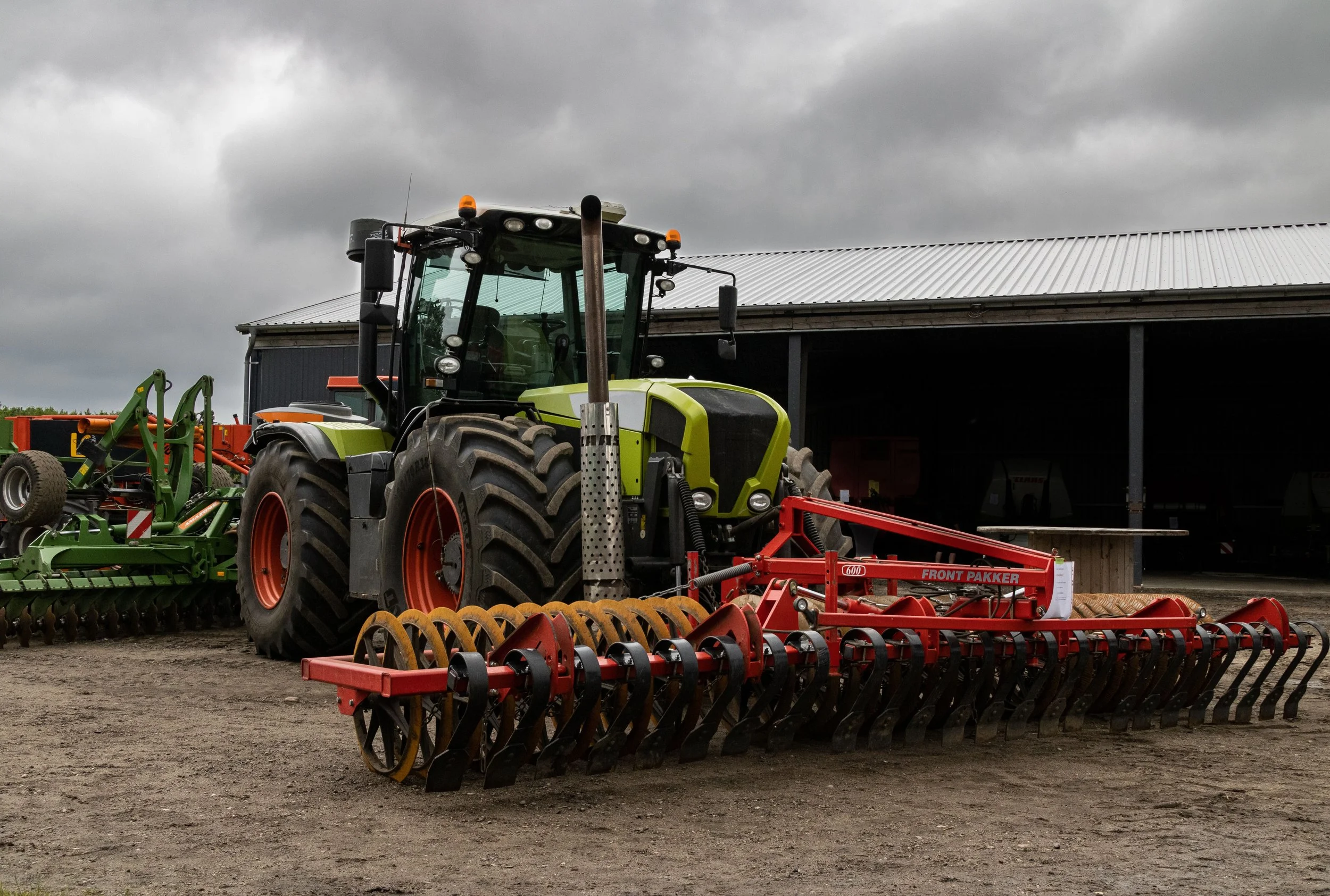 A green tractor attached to red farm equipment on a dirt farm yard with a gray clouded sky and farm buildings in the background.