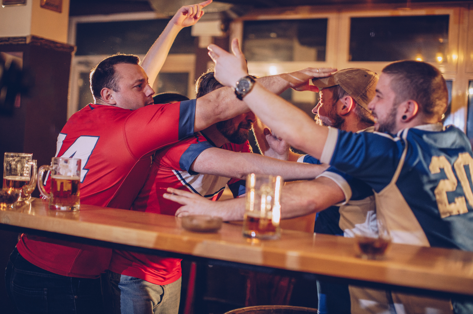 A group of men wearing red and blue sports jerseys fighting in a bar with mugs on the bar top.