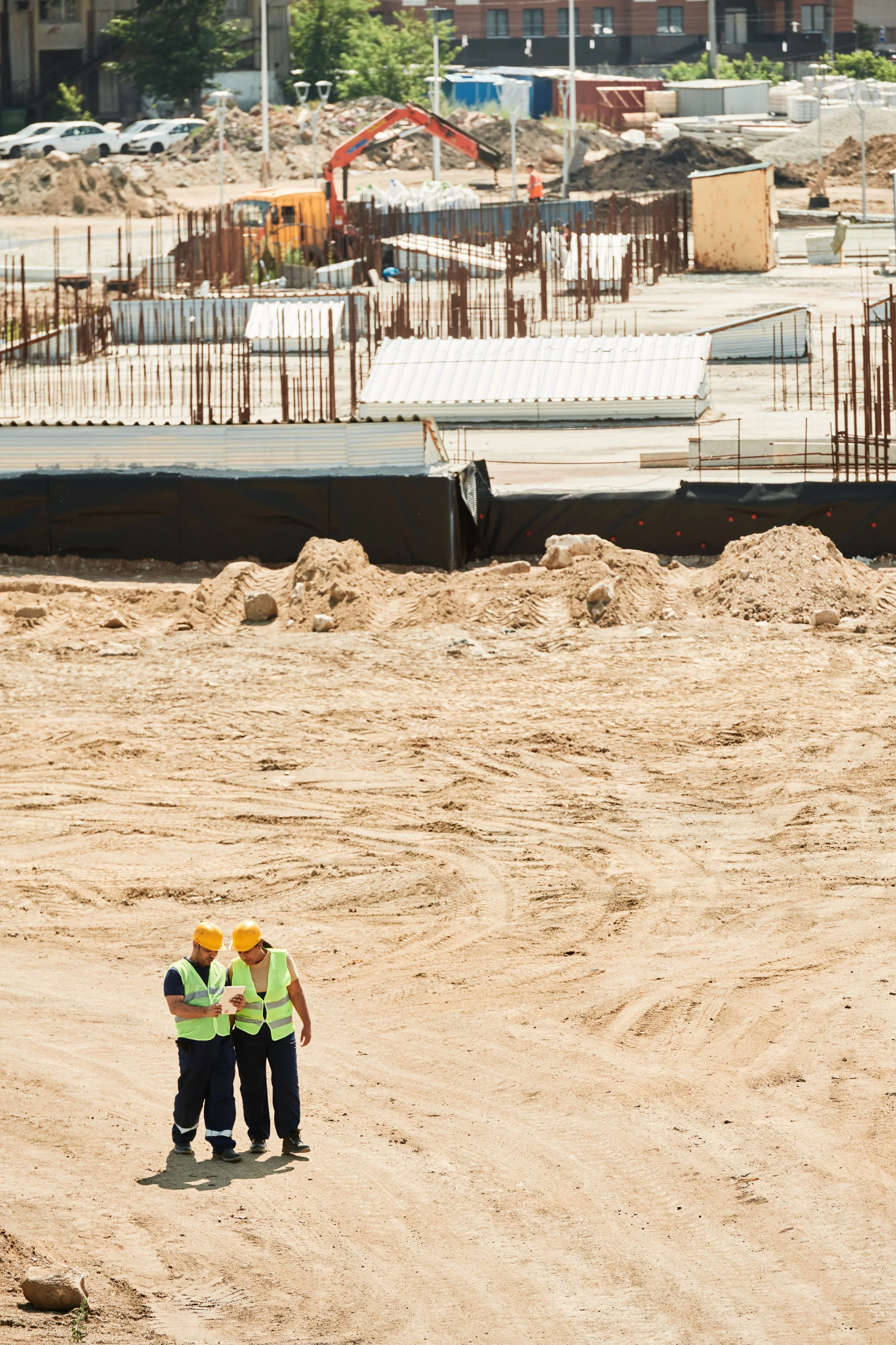 Two construction workers wearing yellow hard hats and green safety vests standing on dirt at a construction site, looking at a tablet. In the background, there is construction equipment, dirt piles, and building materials.