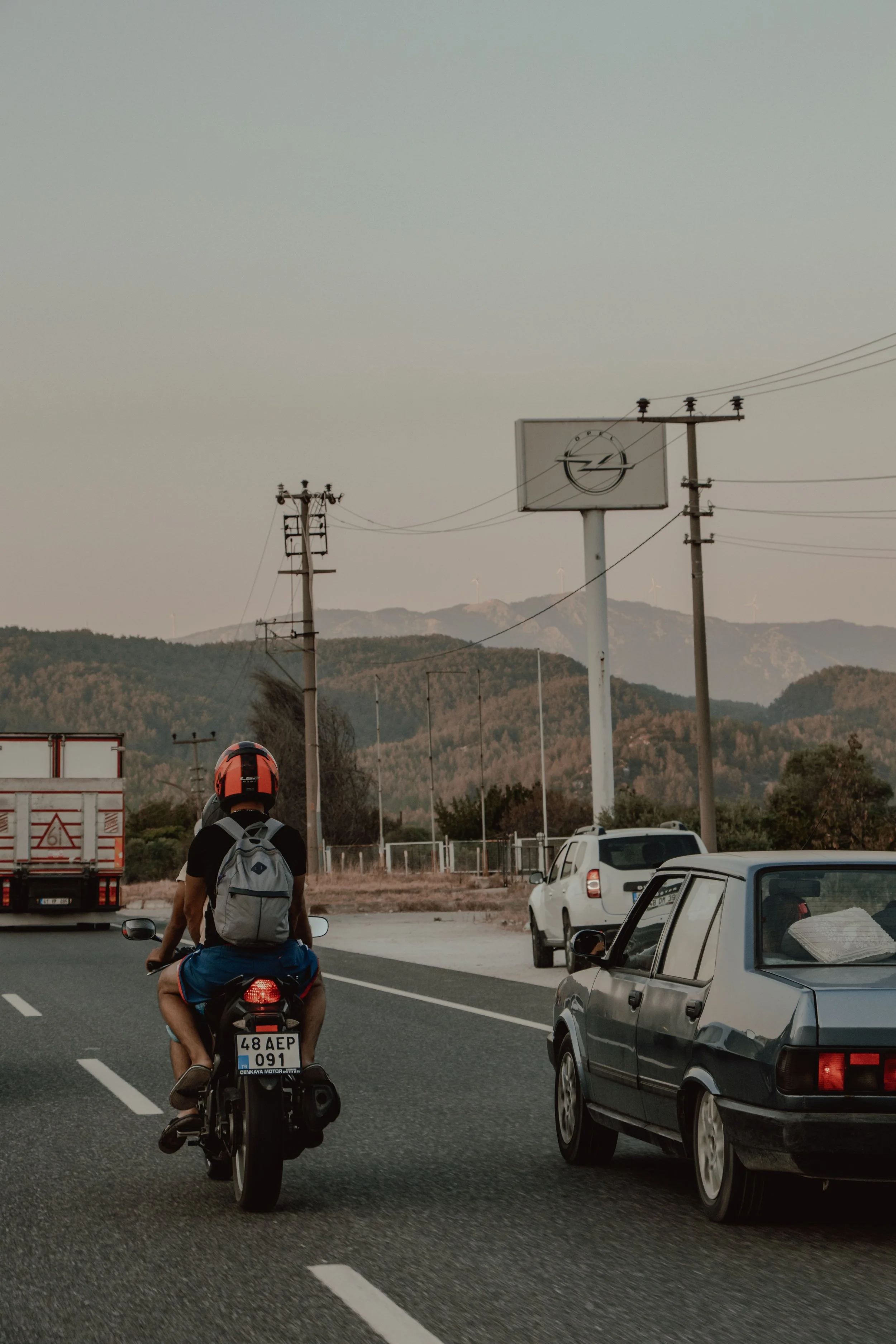 A person riding a motorcycle with a backpack on a busy road, surrounded by parked cars, utility poles, and a large billboard with an Opel logo. Mountains are visible in the background.
