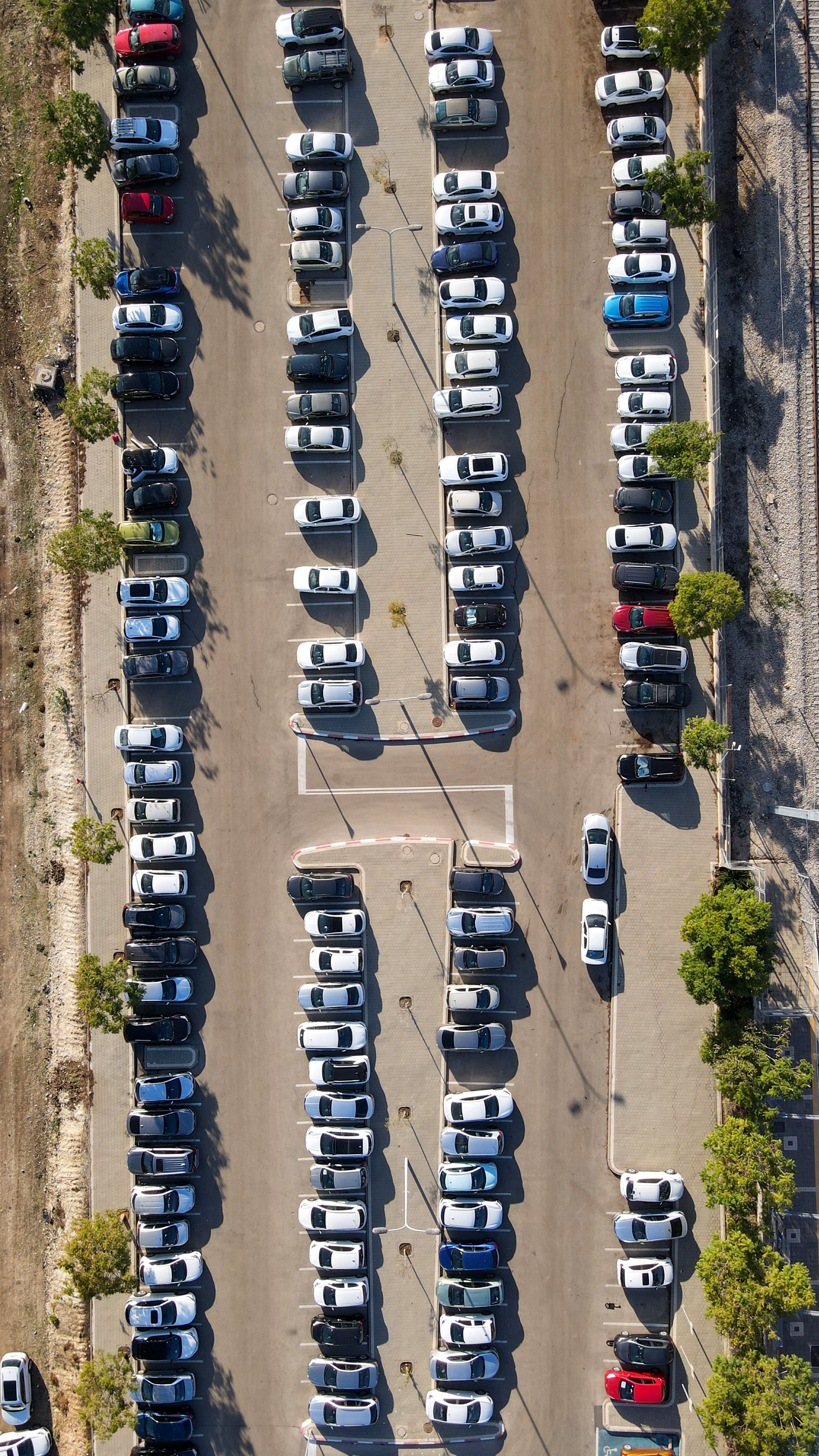 An aerial view of a parking lot with rows of parked cars, some trees lining the lot, and a few empty parking spaces. The cars are mostly white, with a few in other colors.