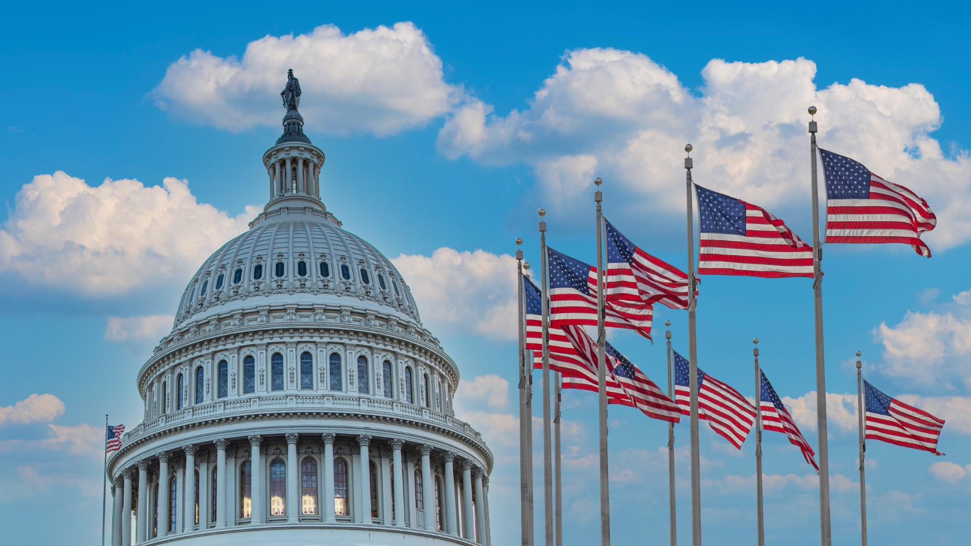 The United States Capitol building with its dome and a row of American flags flying in front on a partly cloudy sky.
