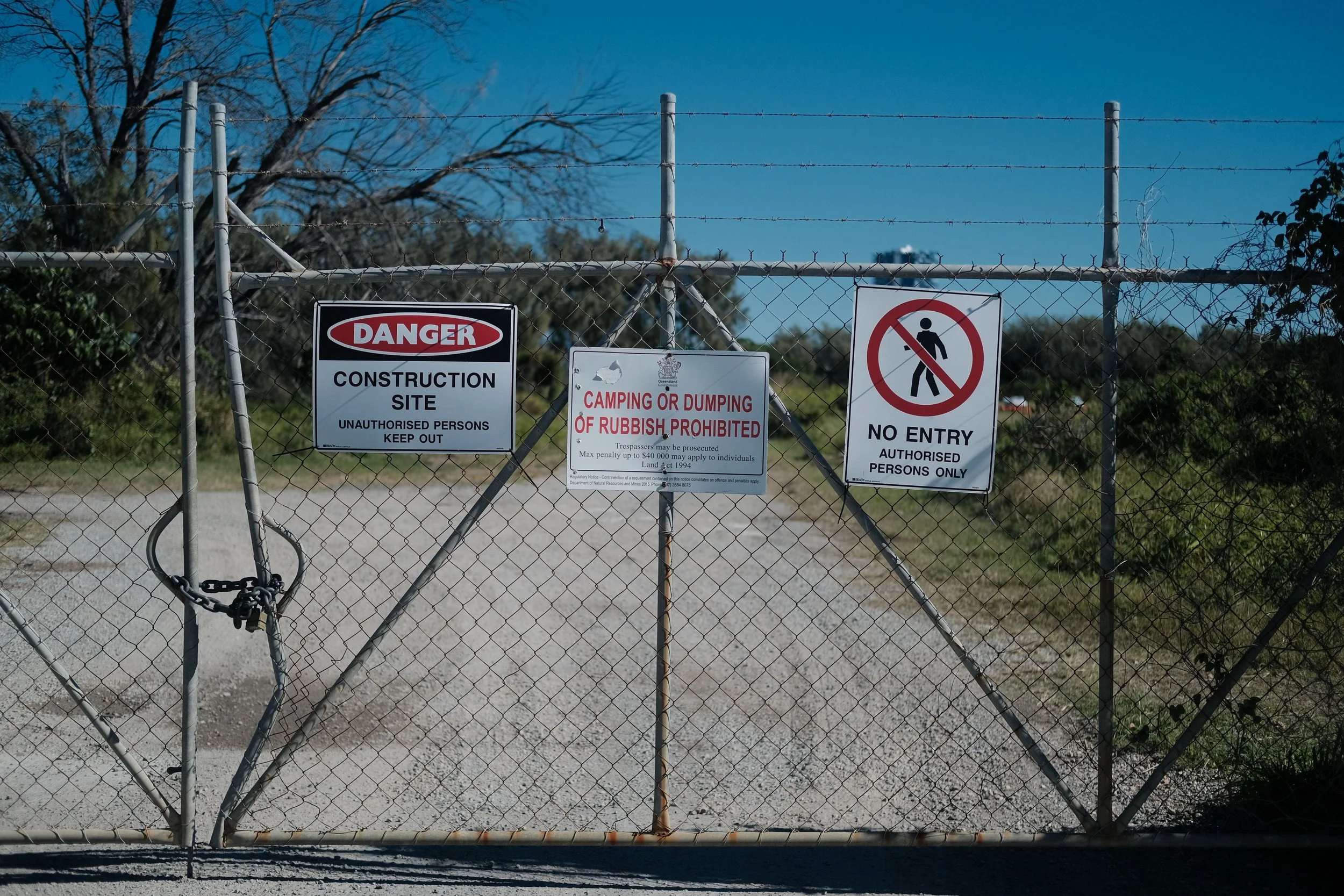 A chain-link fence with multiple warning and prohibitory signs, including a 'Danger Construction Site Keep Out,' 'Camping or Dumping of Rubbish Prohibited,' and 'No Entry Authorized Persons Only,' with a background of trees and a blue sky.