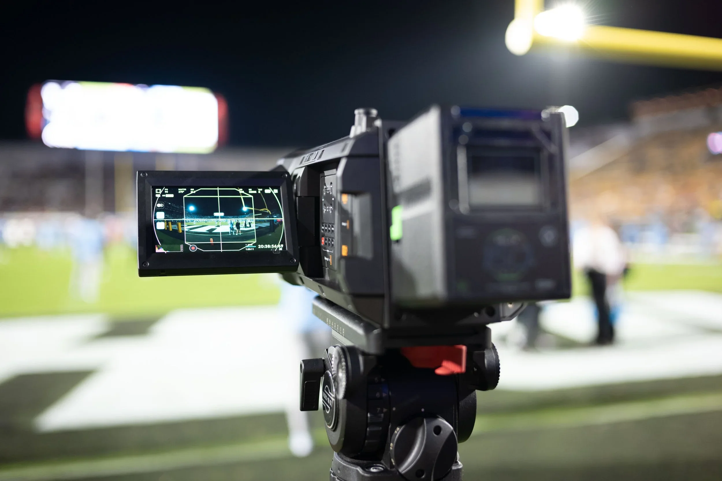 Close-up of the Blackmagic Ursa Cine Immersive video camera on tripod at night, with a brightly lit billboard and stadium lights in the background.