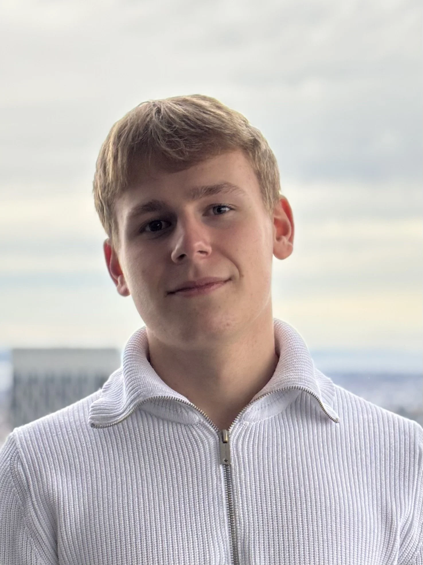 Junge mit dunkelblonden, kurzen Haaren trägt eine weiße, gepunktete Jacke mit Reißverschluss auf einem Balkon mit Blick auf Seattle im Hintergrund.