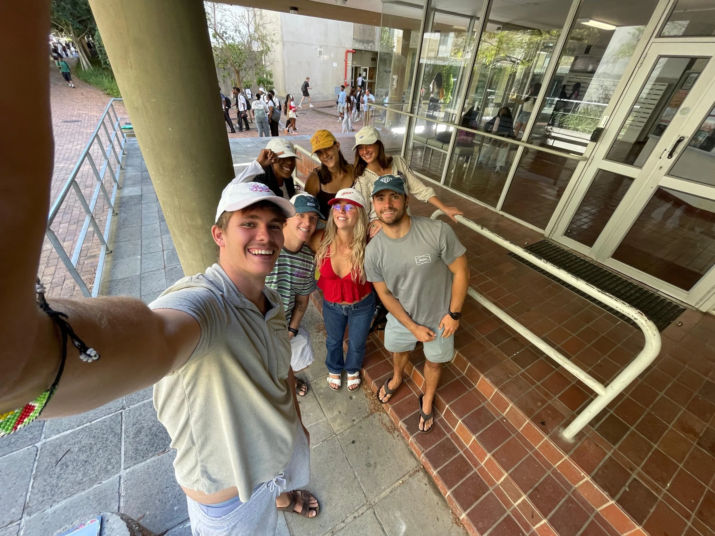 A group of six smiling young adults taking a selfie under a covered walkway outside a building with large glass windows. In the background, several people are walking on a brick-paved path.