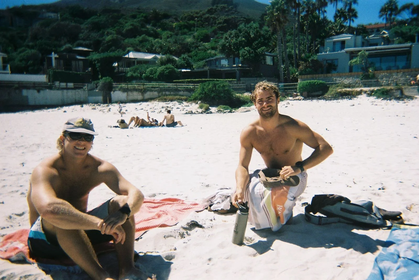 Two men sitting on the sandy beach, smiling at the camera, with beach gear and water bottles nearby. One is wearing a hat and sunglasses, the other is shirtless, with houses and other beachgoers in the background.