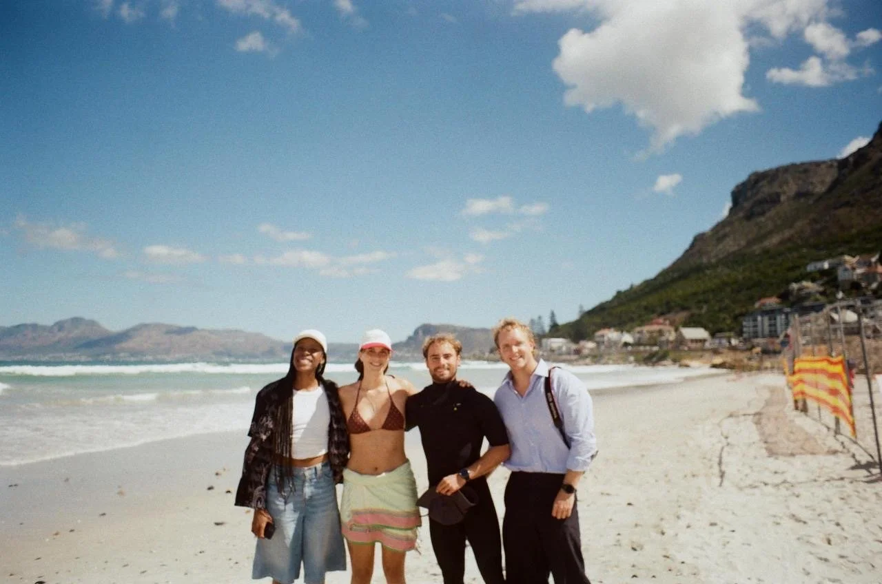 Four people standing on a beach with ocean waves, mountains, and a cloudy sky in the background. They are smiling and dressed in casual summer clothes.