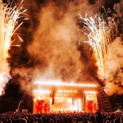 Large outdoor concert with fireworks in the night sky and a crowd watching a stage with performers.
