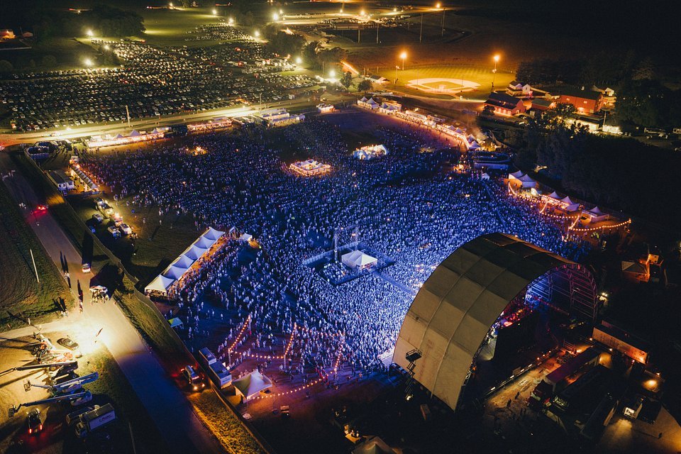 Nighttime aerial view of a large outdoor concert with a crowd, stage, and tents.
