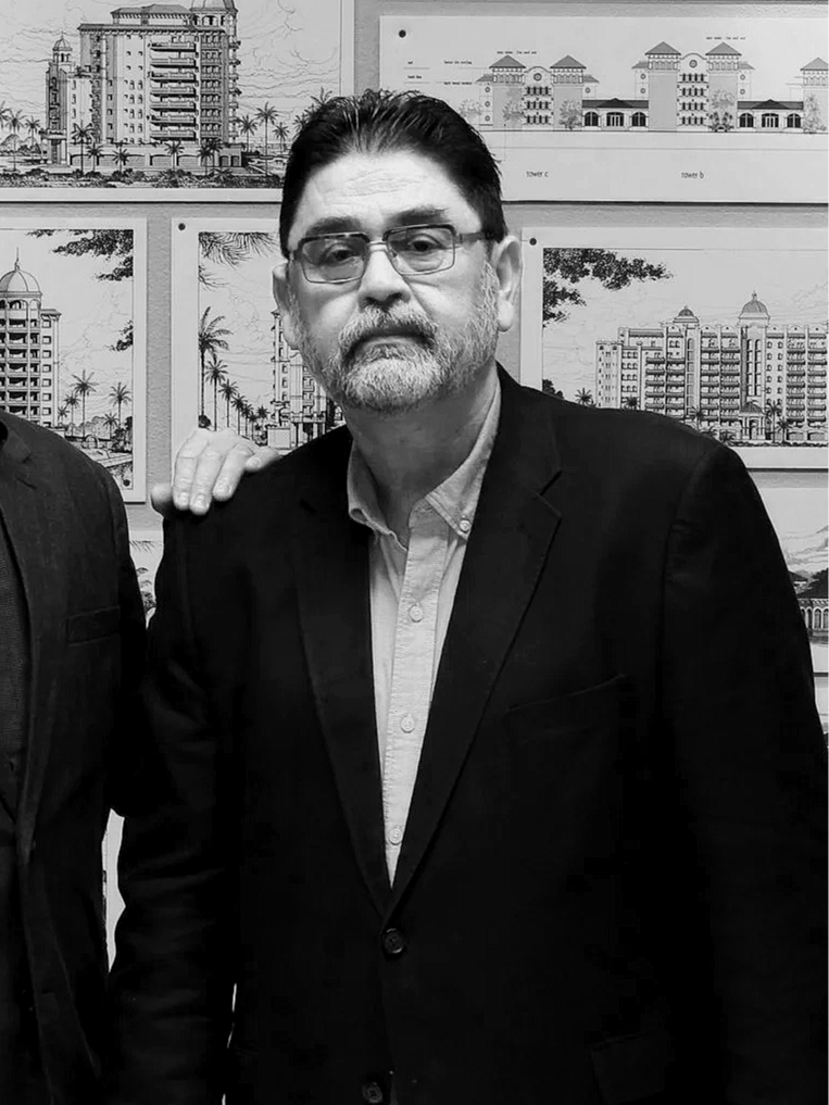 Black and white photo of a middle-aged man with glasses, a beard, and dark hair, wearing a suit jacket and a collared shirt, standing in front of architectural drawings of buildings and towers.