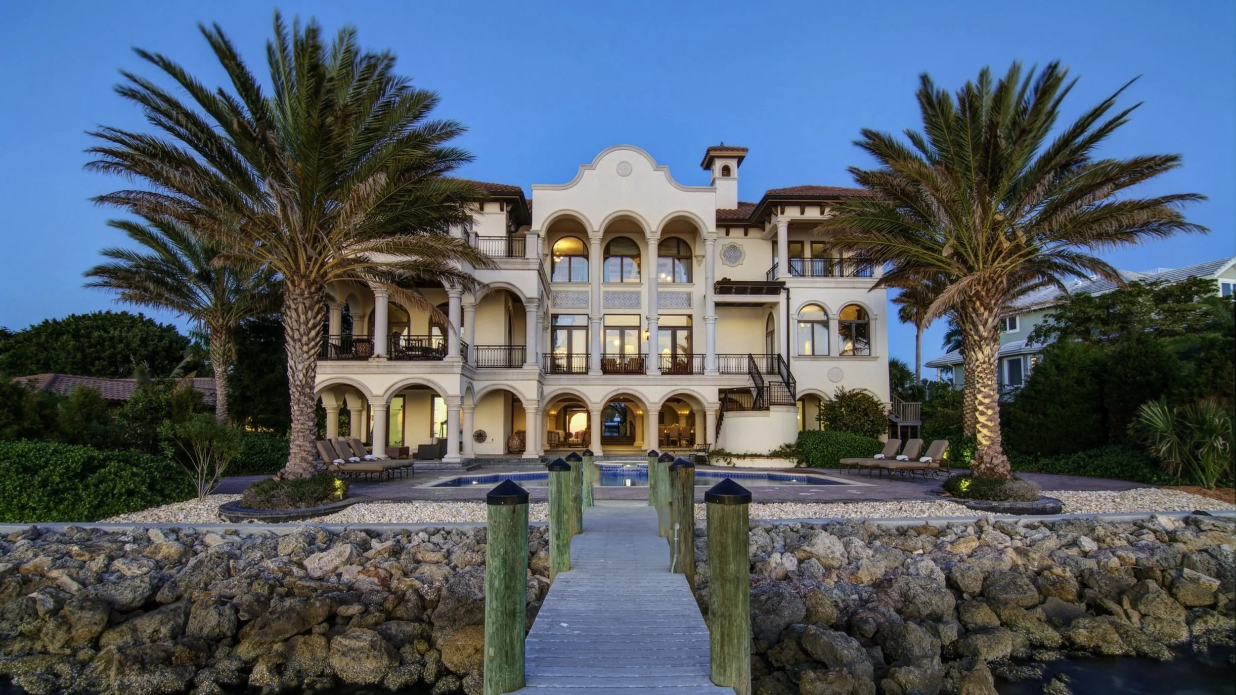 Large white Mediterranean-style house with multiple balconies and arched windows, surrounded by palm trees, with a swimming pool and a wooden dock in the foreground, during twilight.