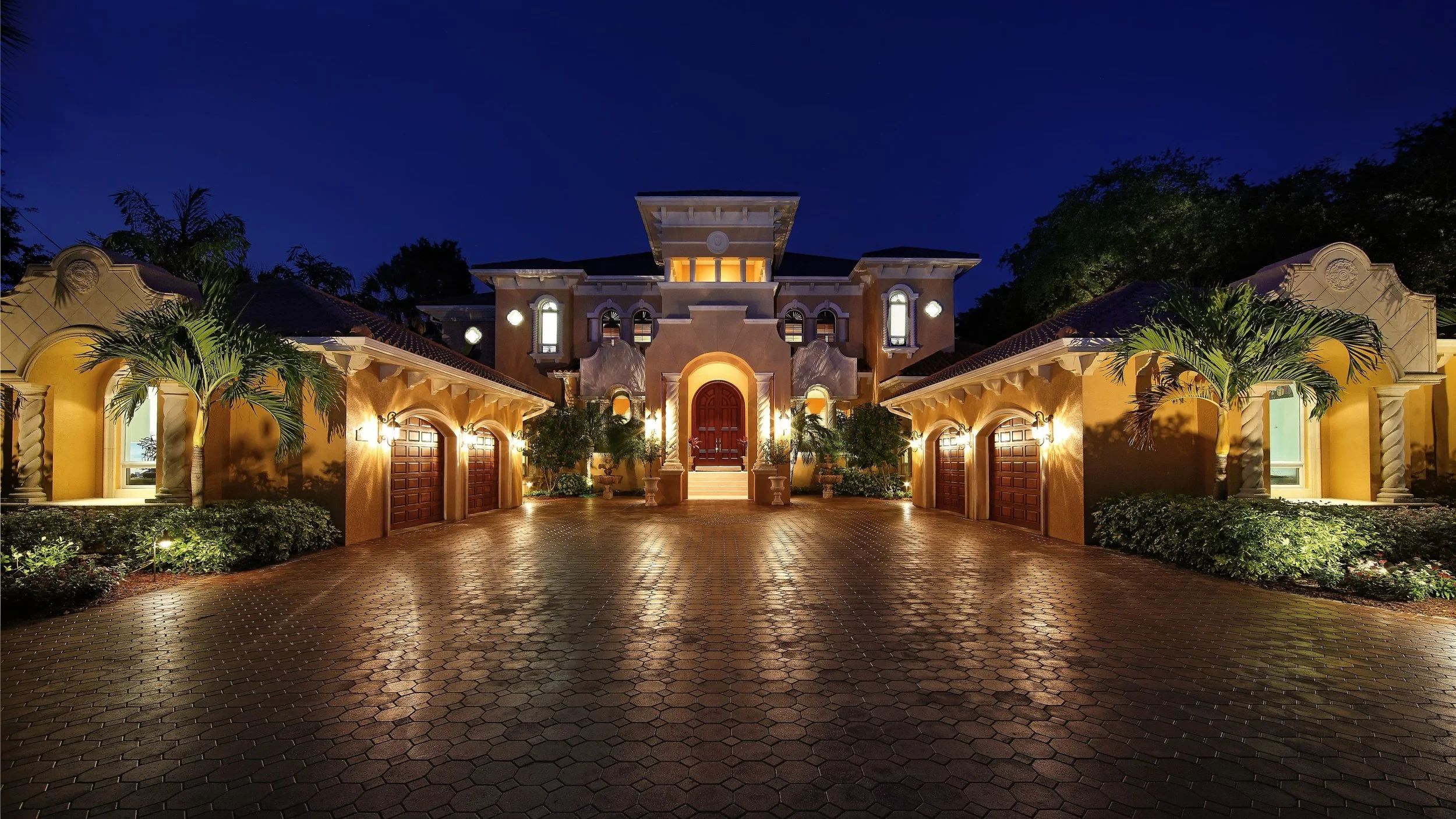 A large, luxurious house illuminated at night with exterior lights, tall palm trees, and a brick driveway.