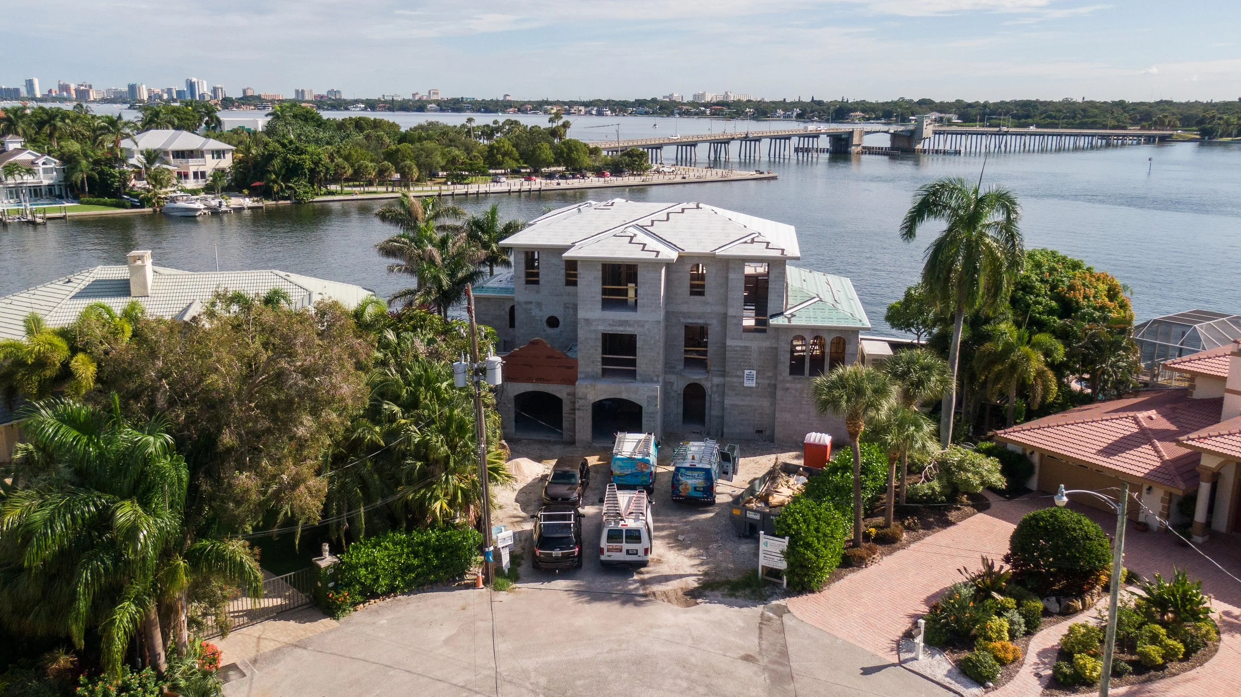 Construction site with a partially built house surrounded by palm trees and other houses, with a waterway and bridge in the background.