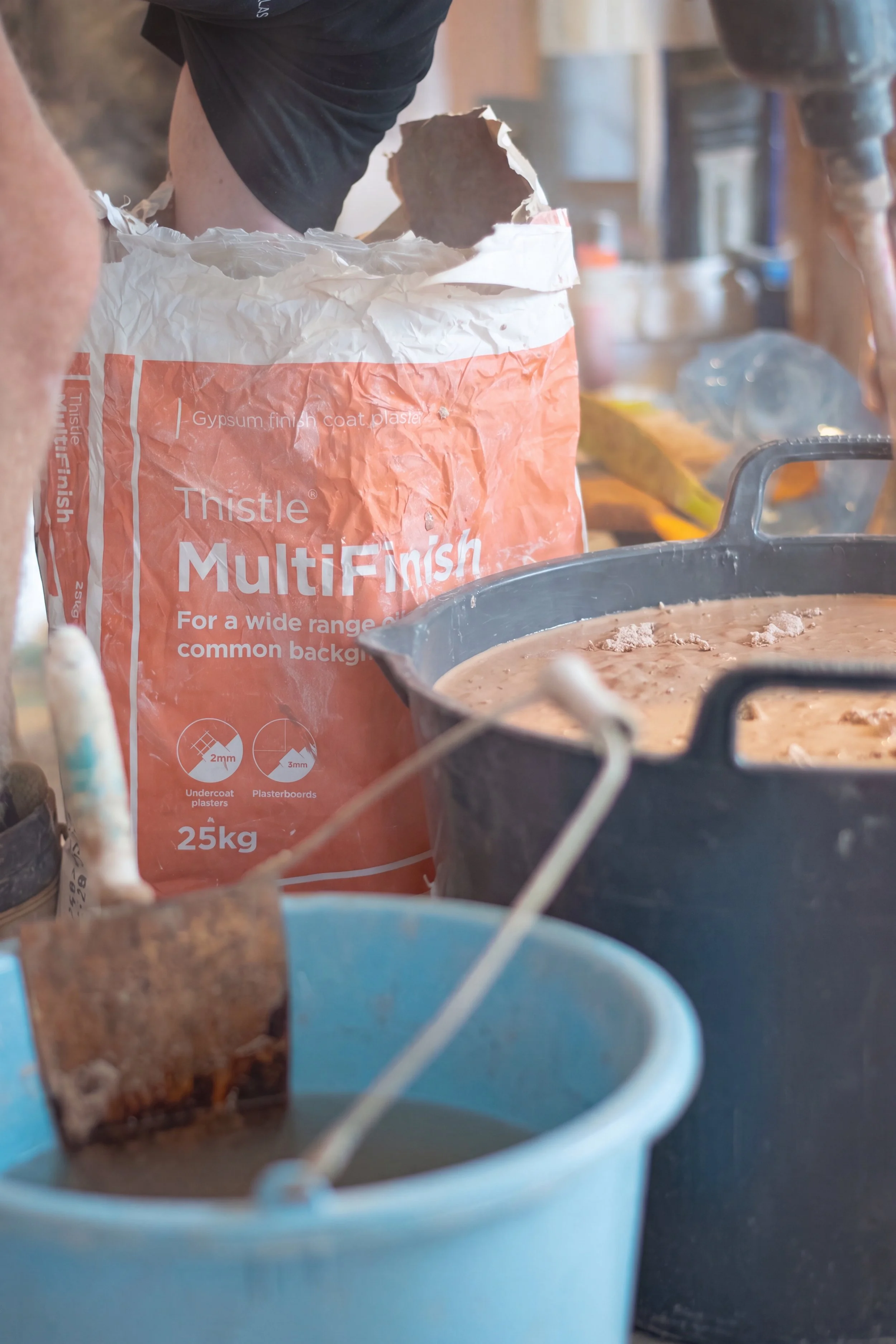 Close-up of a partially open bag of Thistle MultiFinish plaster with a small hole in the top, surrounded by various tools and containers on a workbench in a workshop setting.