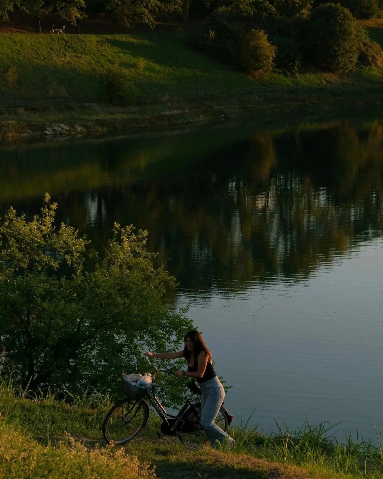 A woman with long dark hair, wearing a black tank top and light-colored pants, is standing next to a bicycle by a lake at sunset. She is smiling and is near a green bush and tall grass, with the calm water and green trees reflected in the lake behind her.