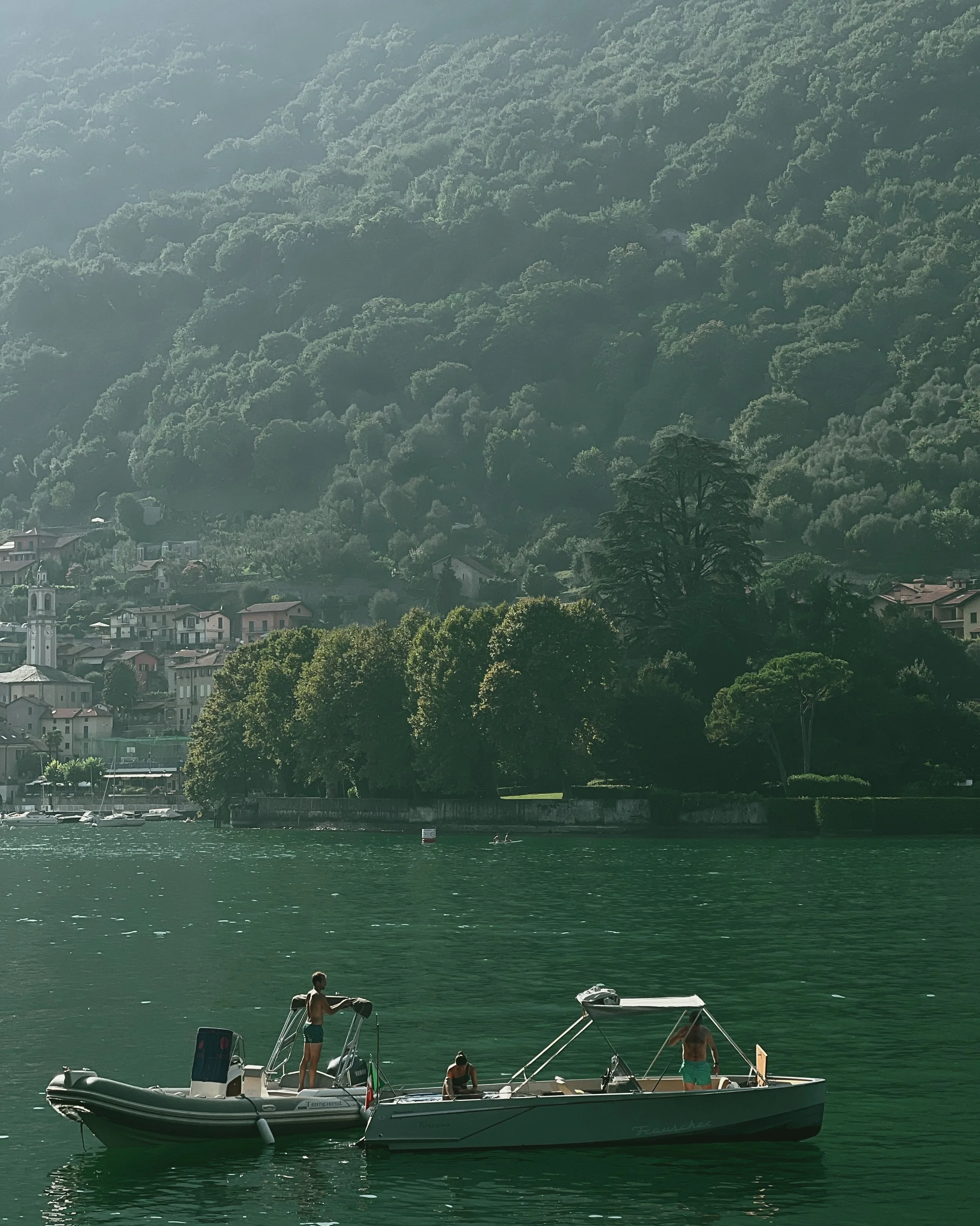 People on boats in a body of water with a hillside covered in trees and houses in the background.