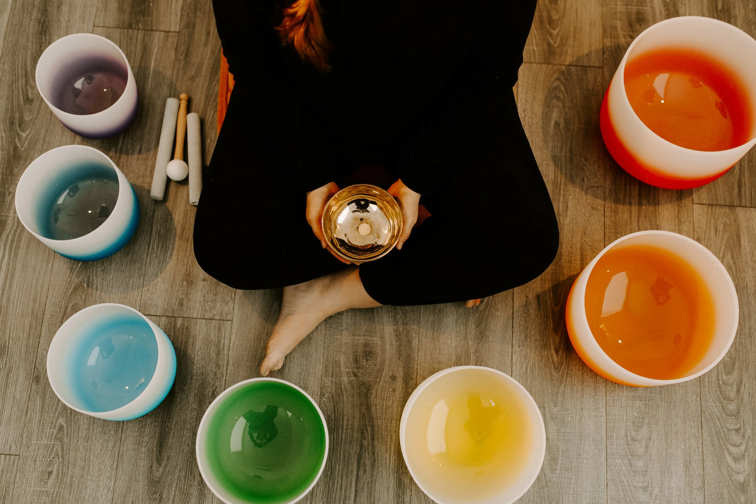 A person sitting on a wooden floor holding a Tibetan bowl, surrounded by colorful crystal singing bowls in shades of purple, blue, green, yellow, orange, and white, with two white and wooden mallets.