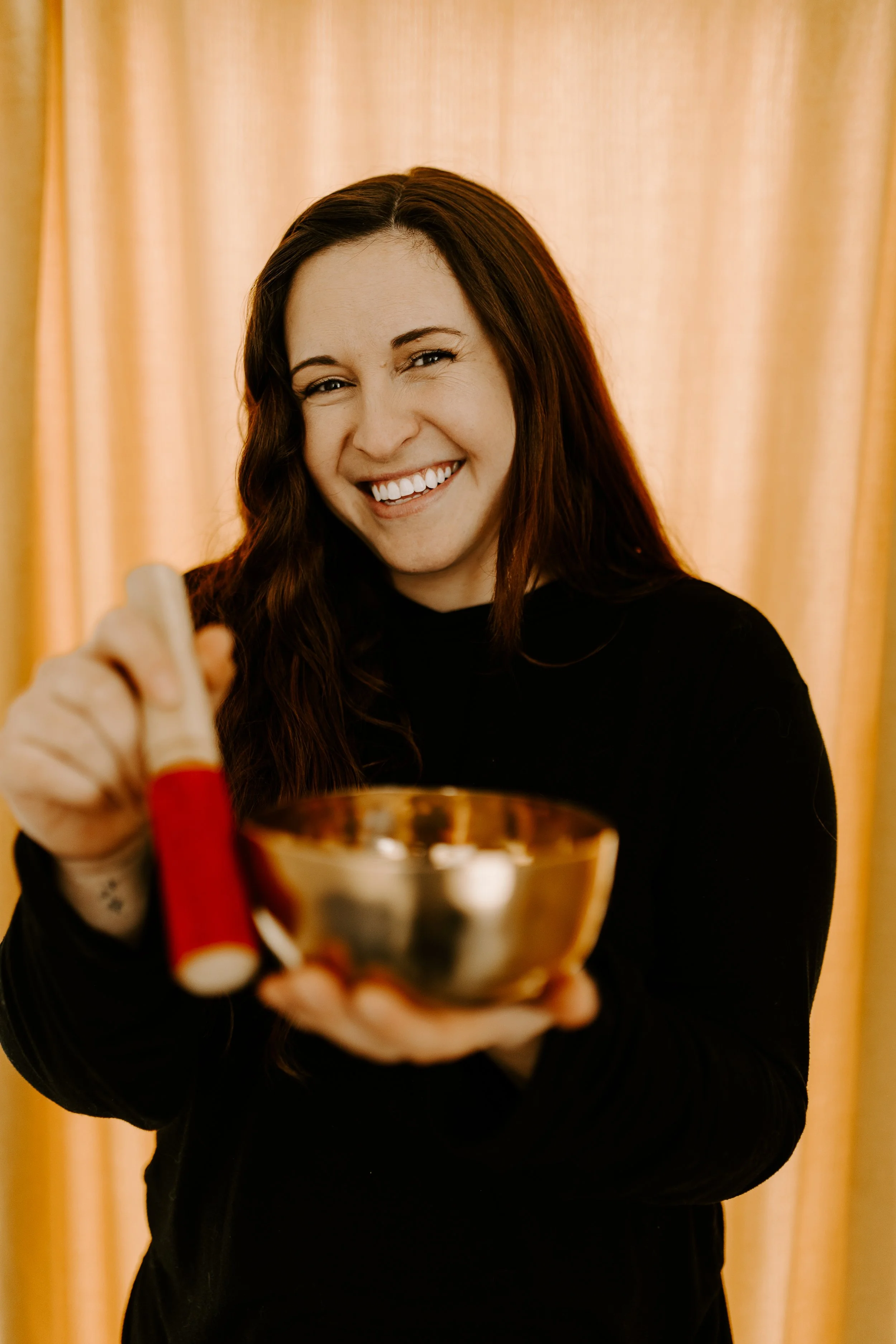 A woman with long brown hair smiling and holding a Tibetan singing bowl in front of a yellow curtain.