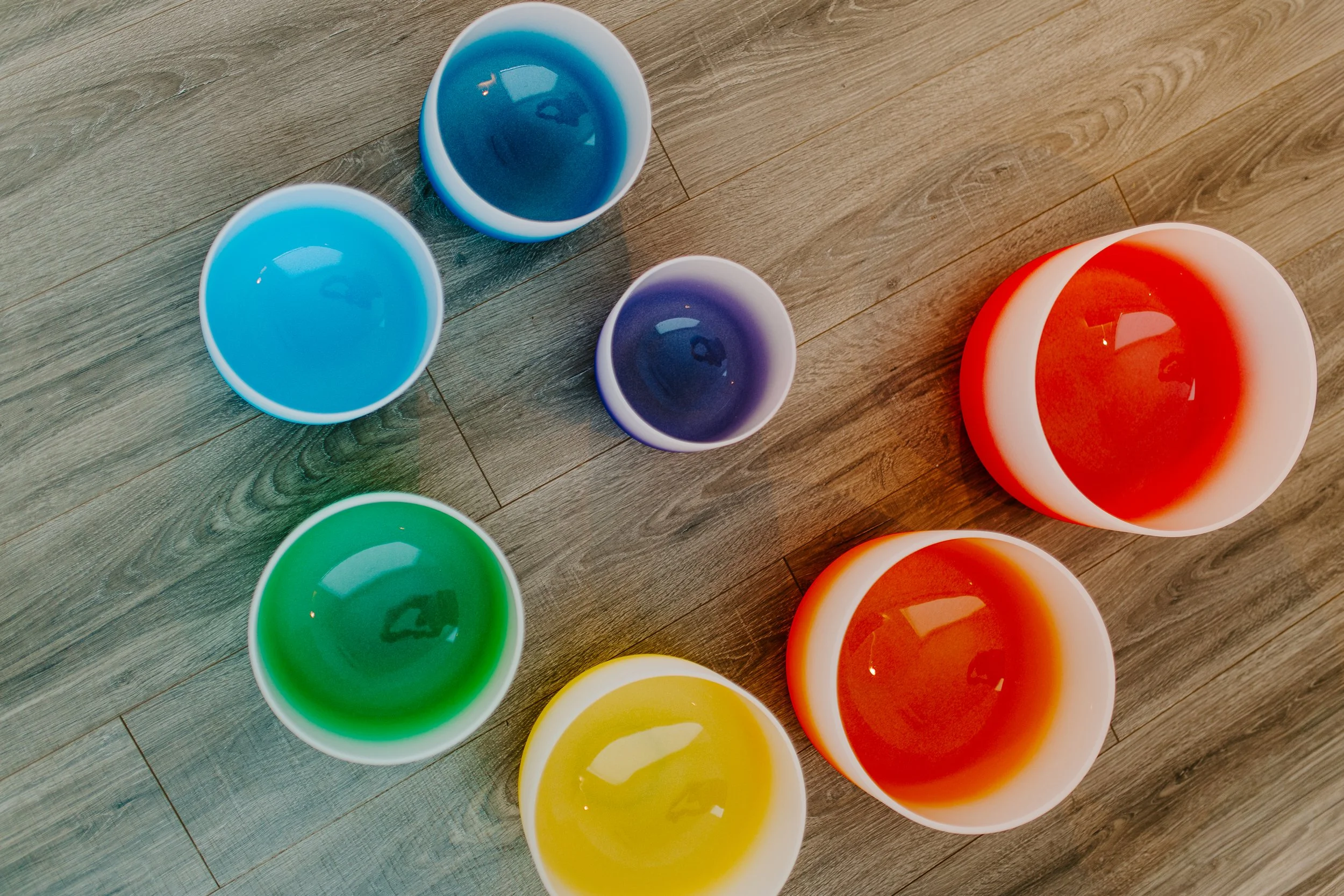 Several colorful crystal singing bowls  arranged on a wooden floor.