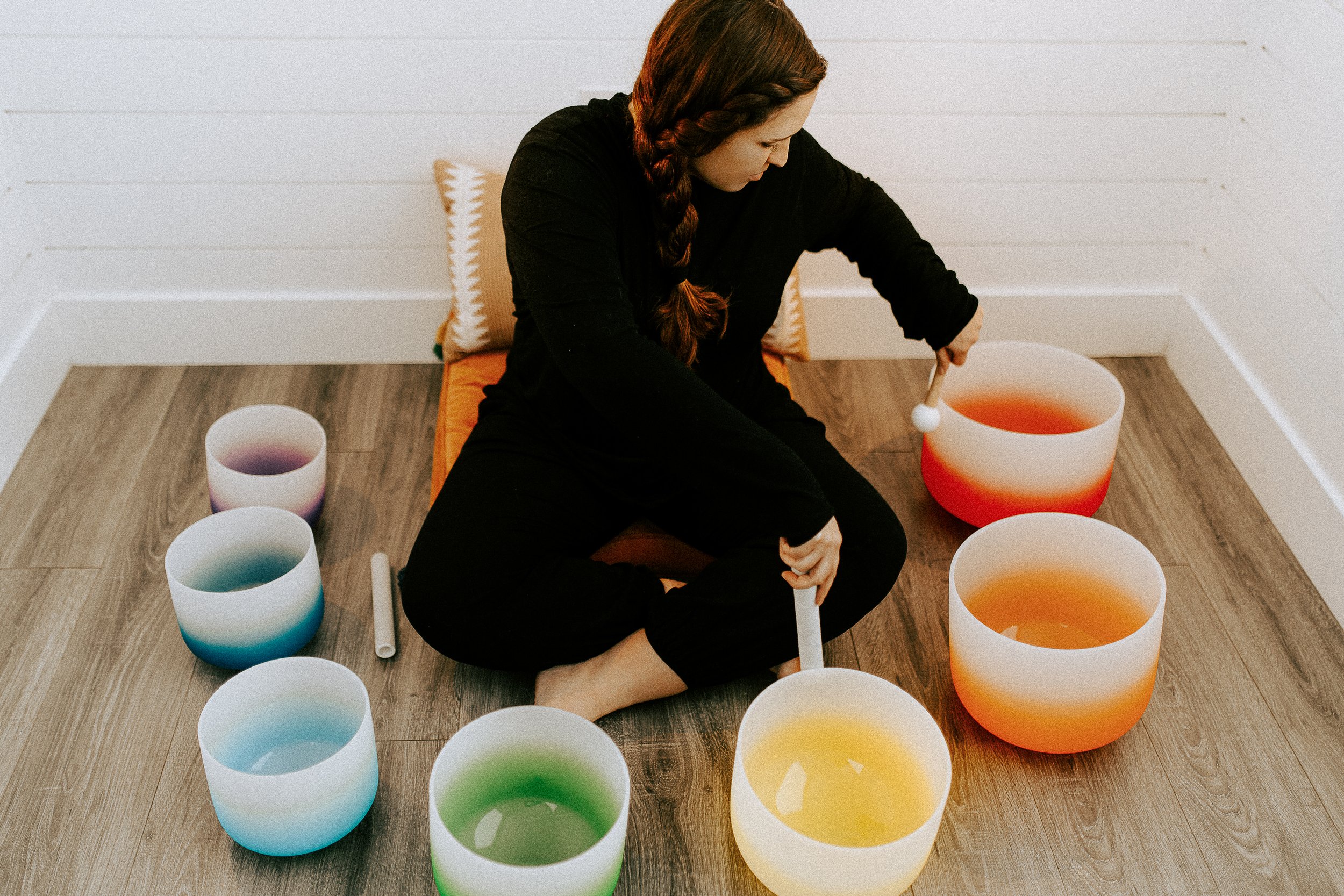 A woman in black clothing sitting on a cushion on a wooden floor, surrounded by colorful singing bowls filled with water, with a mallet in her hand, engaging in sound therapy or meditation.