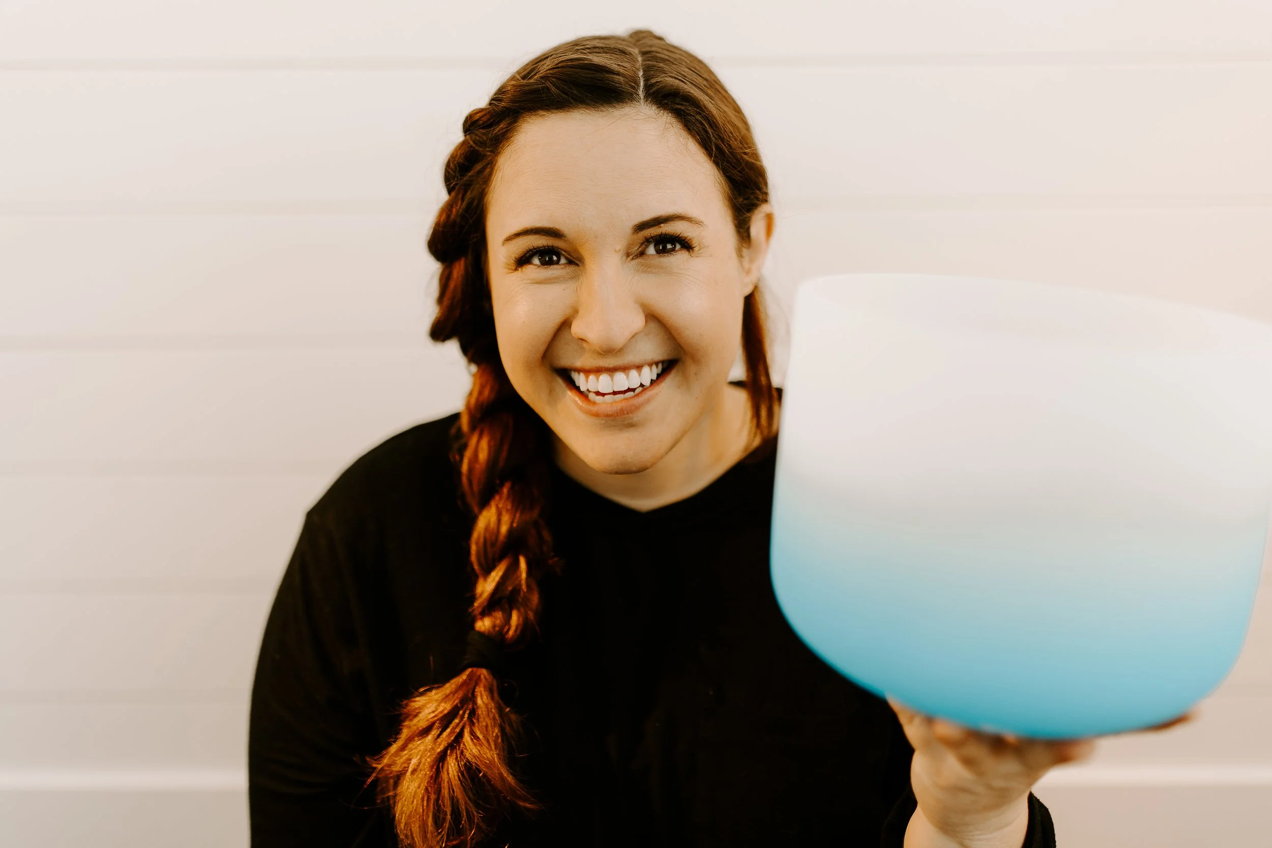 A woman with long brown hair and a braid, smiling while holding a blue crystal singing bowl in front of her