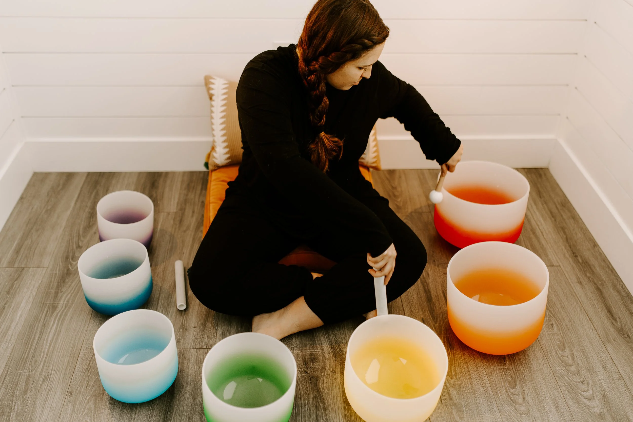 A woman with braided hair is sitting on the floor, playing colorful crystal singing bowls with a mallet in a bright, minimalist room with white walls.
