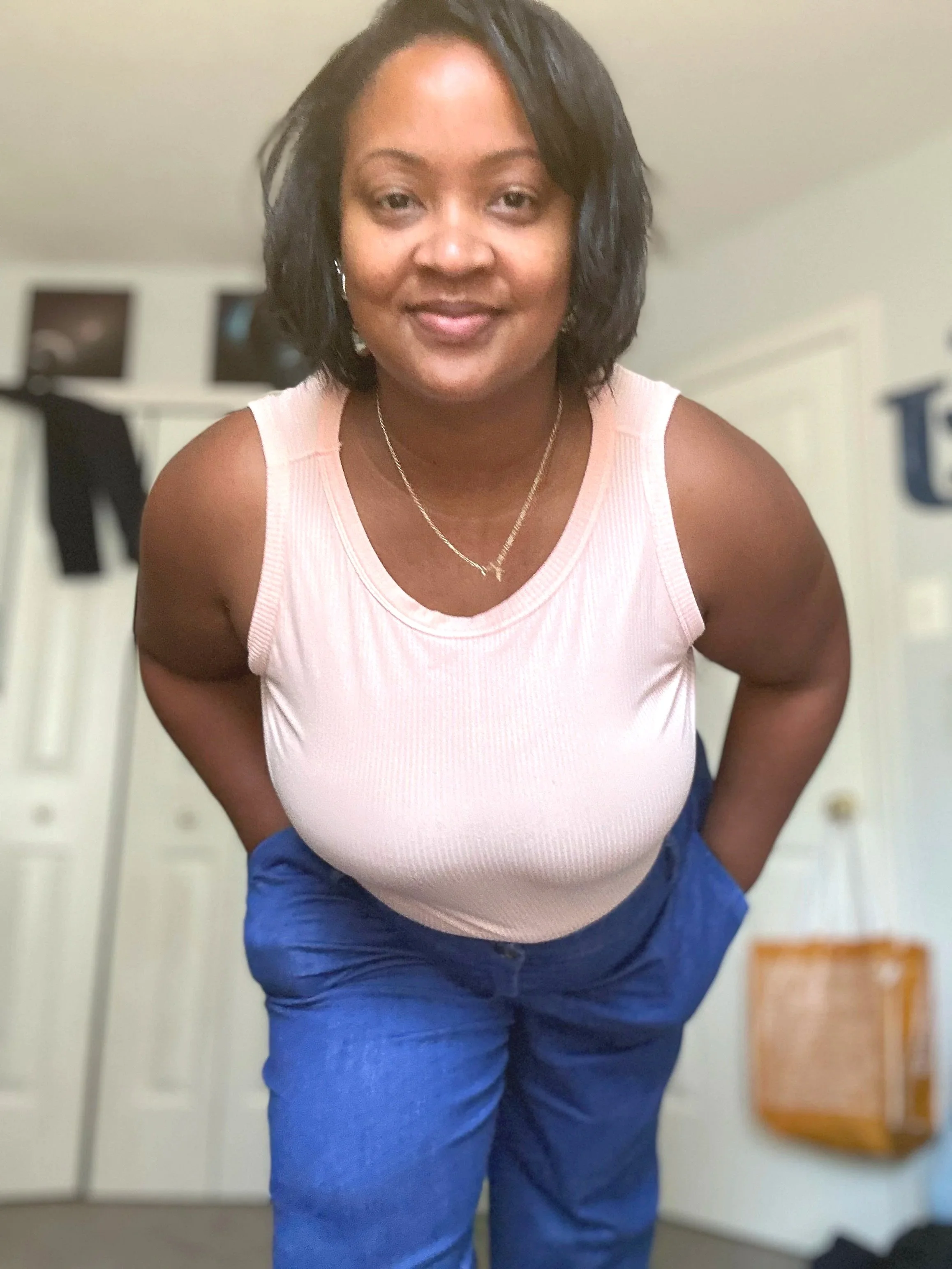 Tajuana, with short curly black hair, wearing a light pink sleeveless top and blue pants, posing for a selfie in front of a mirror, with a blurred background including a white door and a dresser.