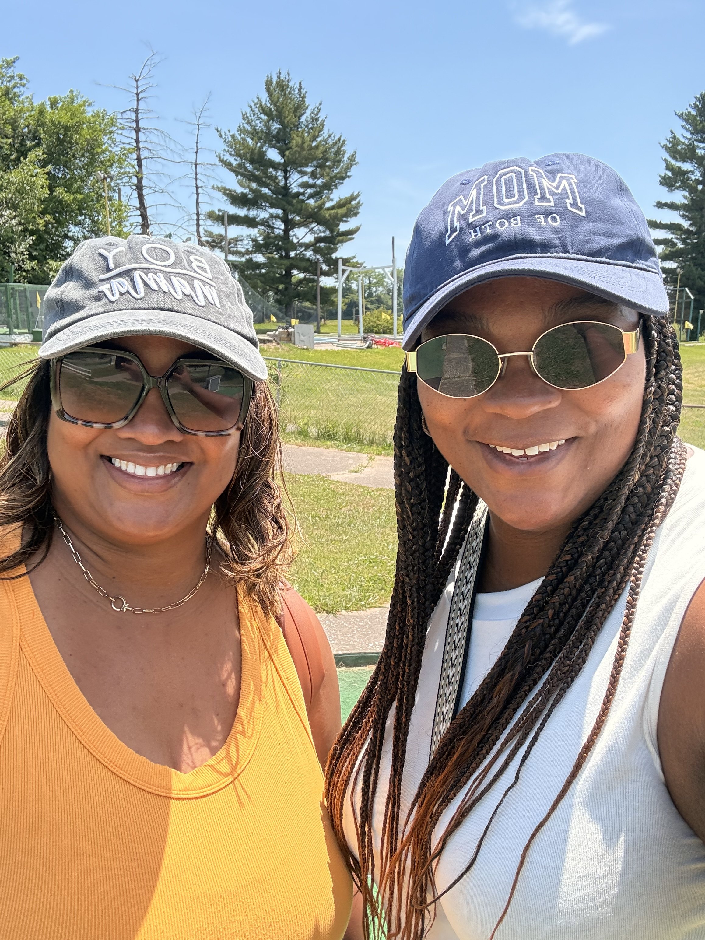 Sisters Tajuana and Sheree are smiling and wearing caps, sunglasses, and casual clothing outdoors on a sunny day with trees in the background.
