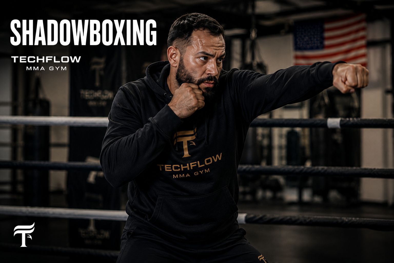A man practicing shadowboxing in a gym, wearing a black hoodie with 'TECHFLOW MMA GYM' logo, in a boxing ring with an American flag in the background.