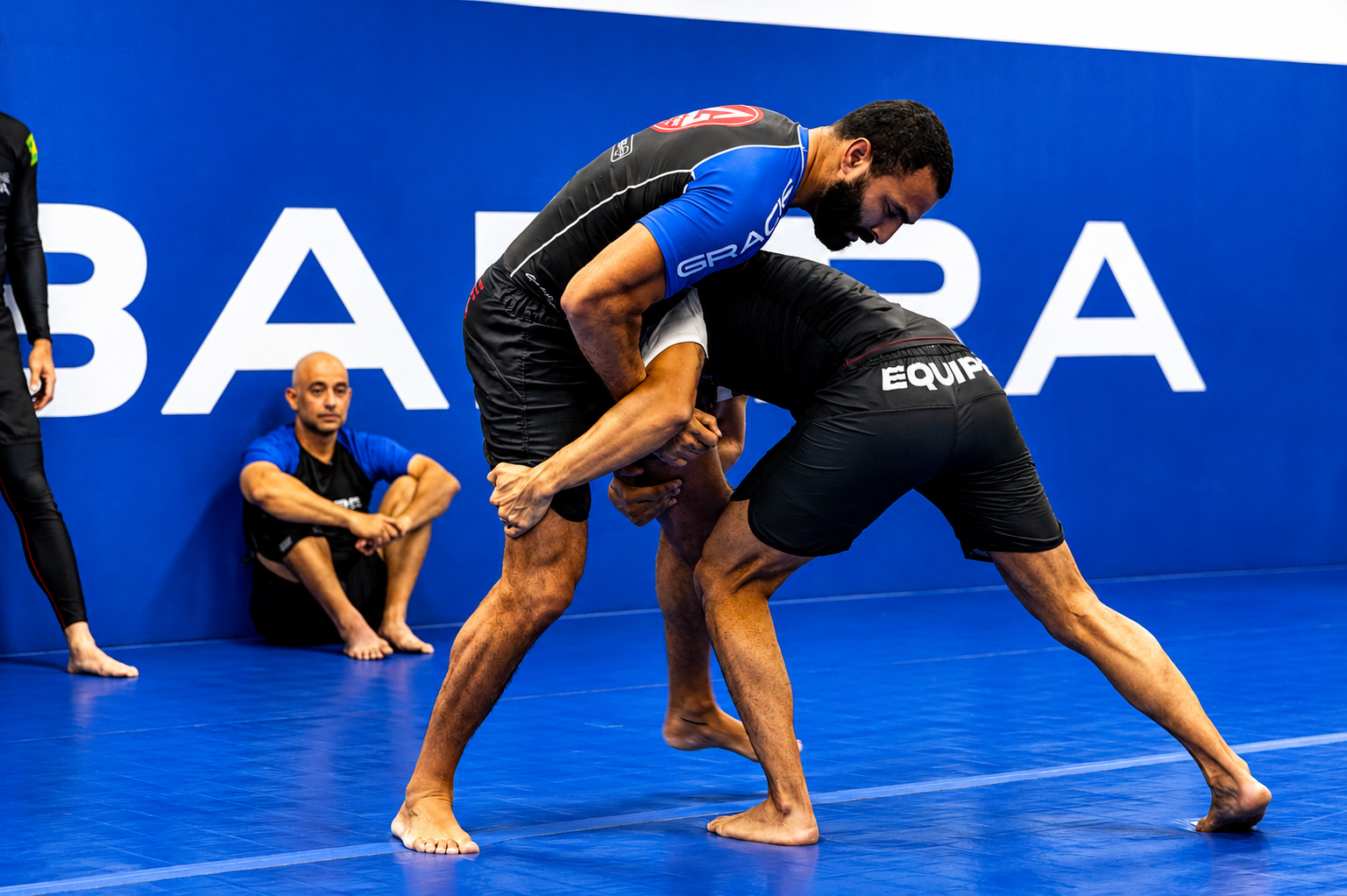 Two men practicing Brazilian Jiu-Jitsu on a blue mat, with one man gripping the other's gi and applying a technique, while a third person sits against the wall watching.