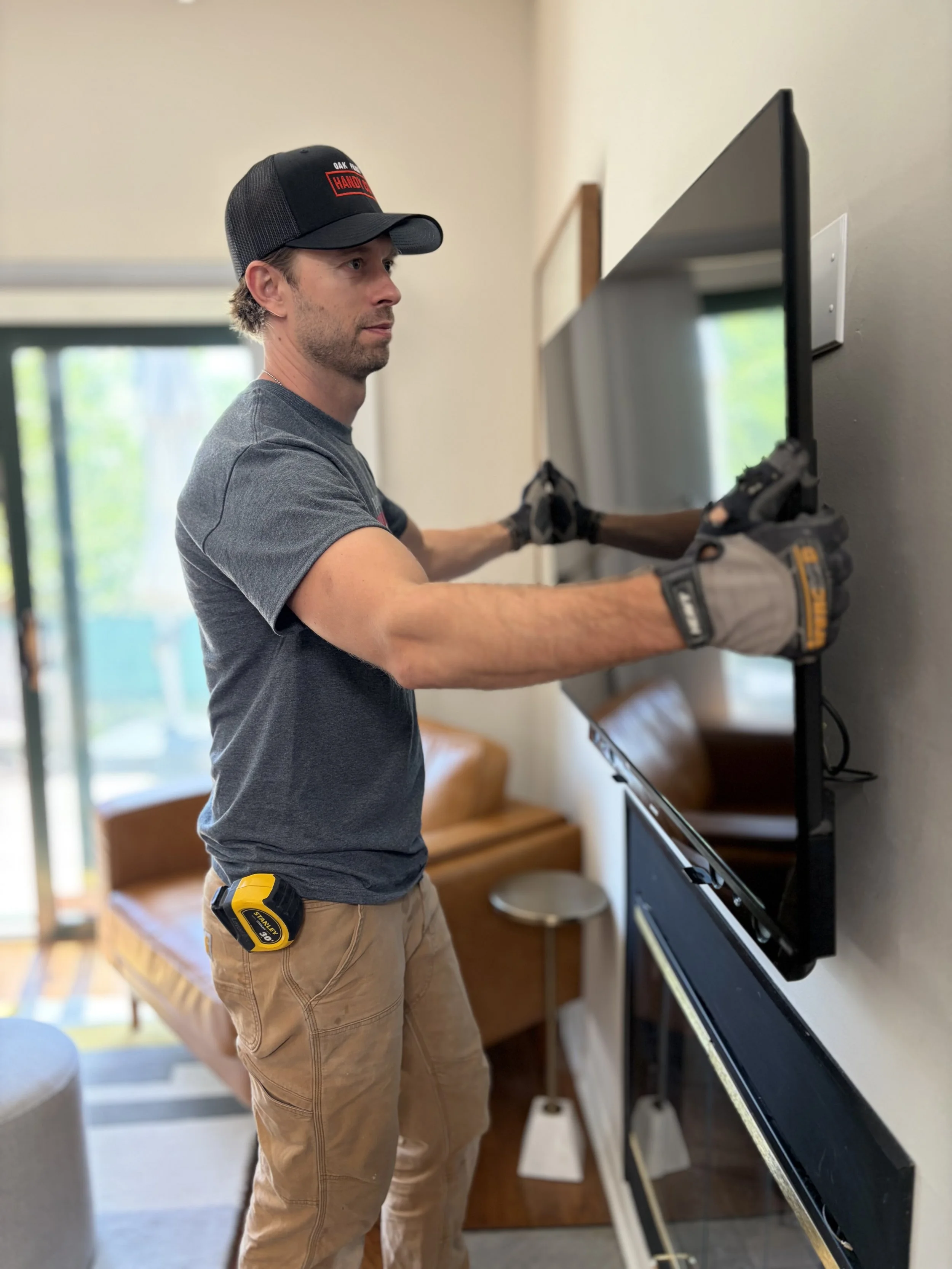 Handyman in a cap and work gloves mounting a flat-screen TV on a wall inside a Forest Park home.