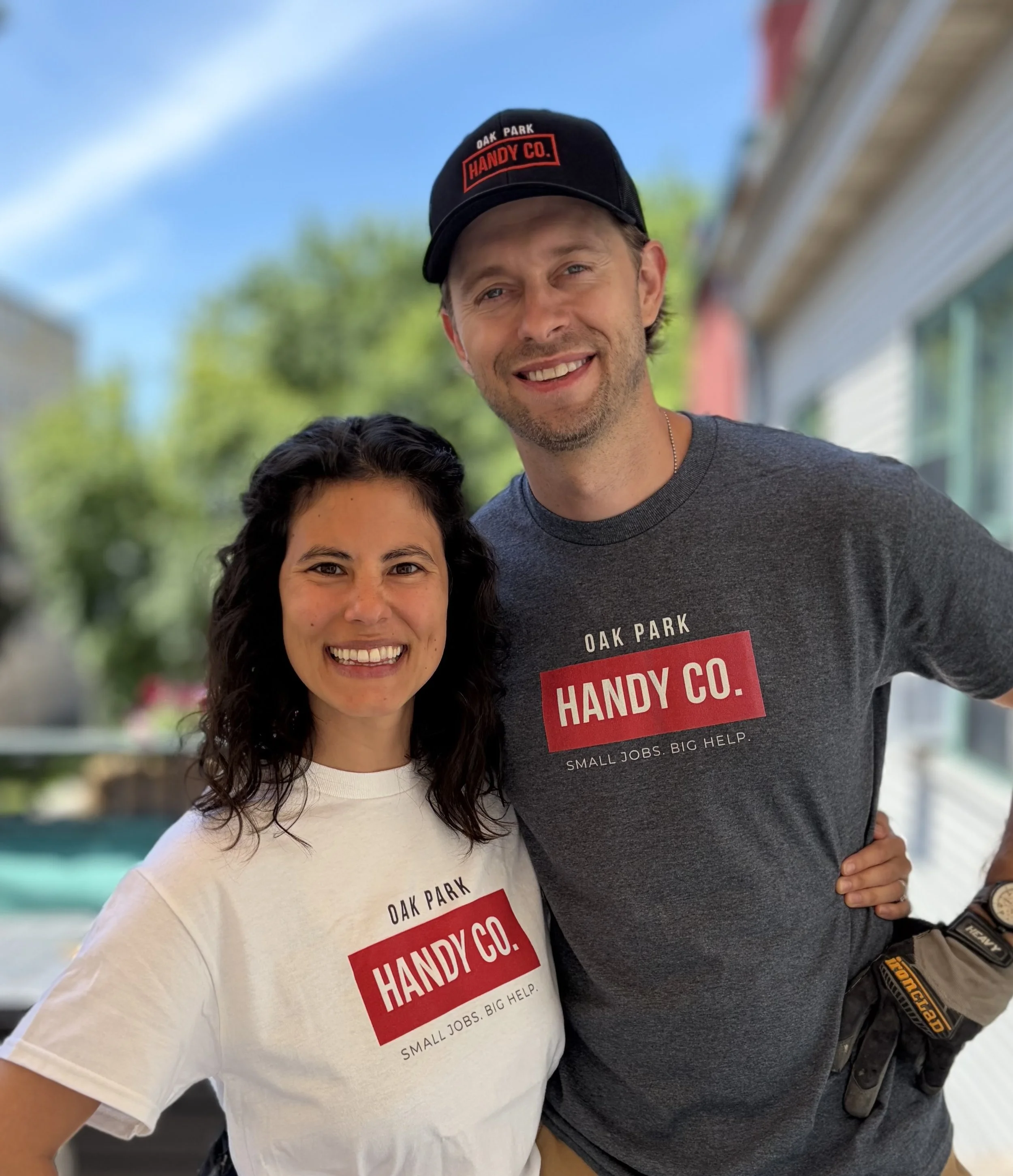 Two smiling people, one male and one female, husband and wife handyman team, wearing Oak Park Handy Co. t-shirts. The man is also wearing a black hat with red text. They are outdoors on a sunny day with trees and buildings in the background.