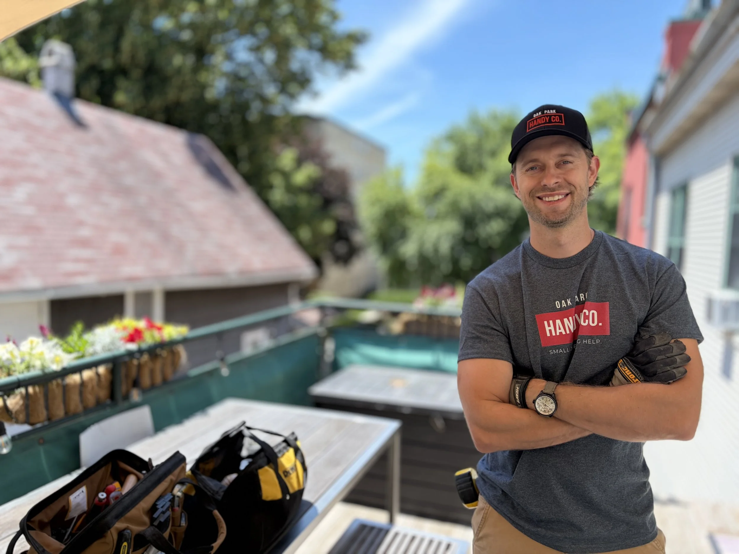 Oak Park handyman smiling wearing an Oak Park Handy Co. branded black hat and gray t-shirt, standing outdoors next to power tools and tool bags, with a house and trees in the background.