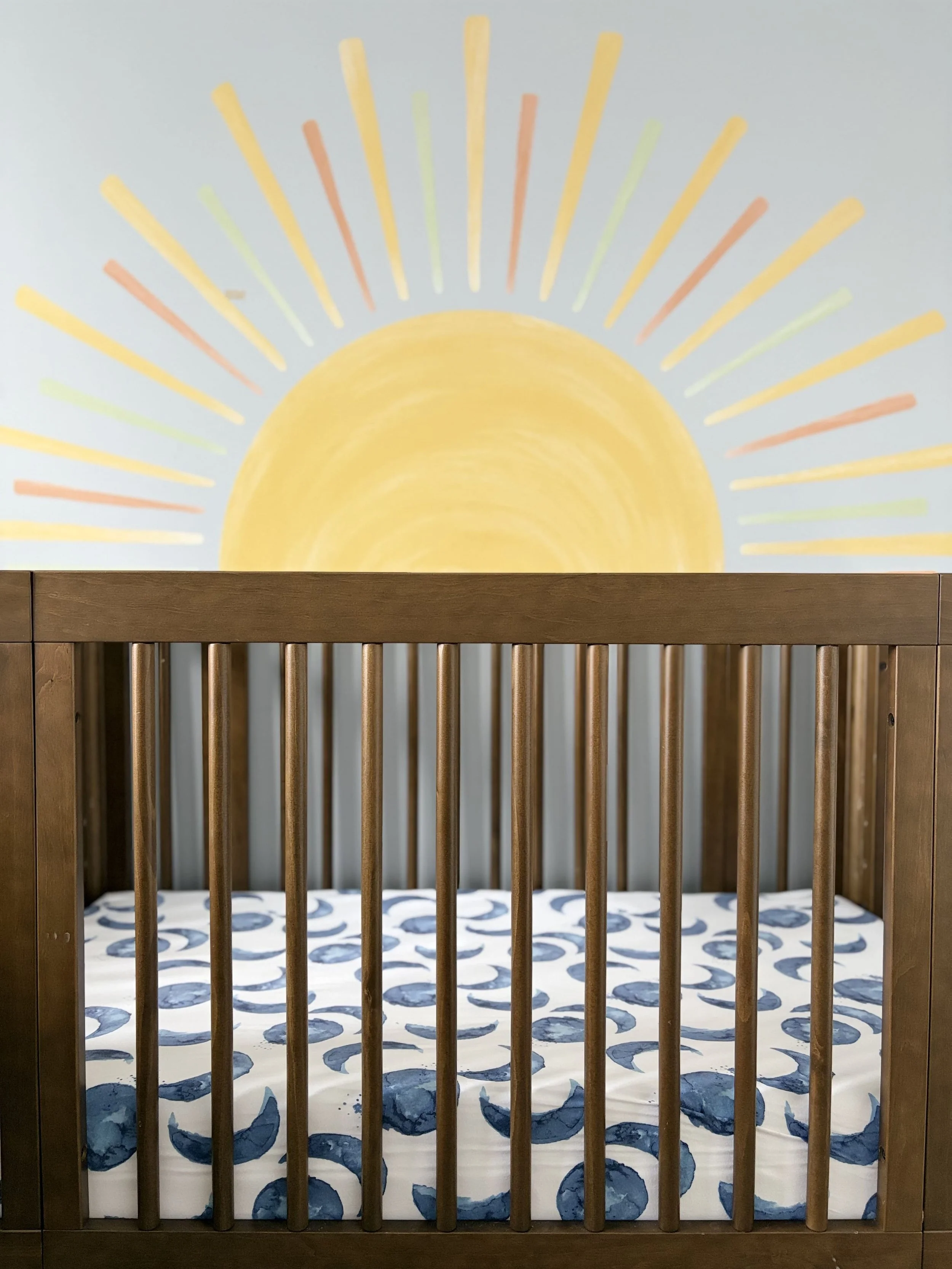 Wooden crib assembled by handyman with a mattress that has a blue and white pattern, and a wall art of a yellow sun with multicolored rays.