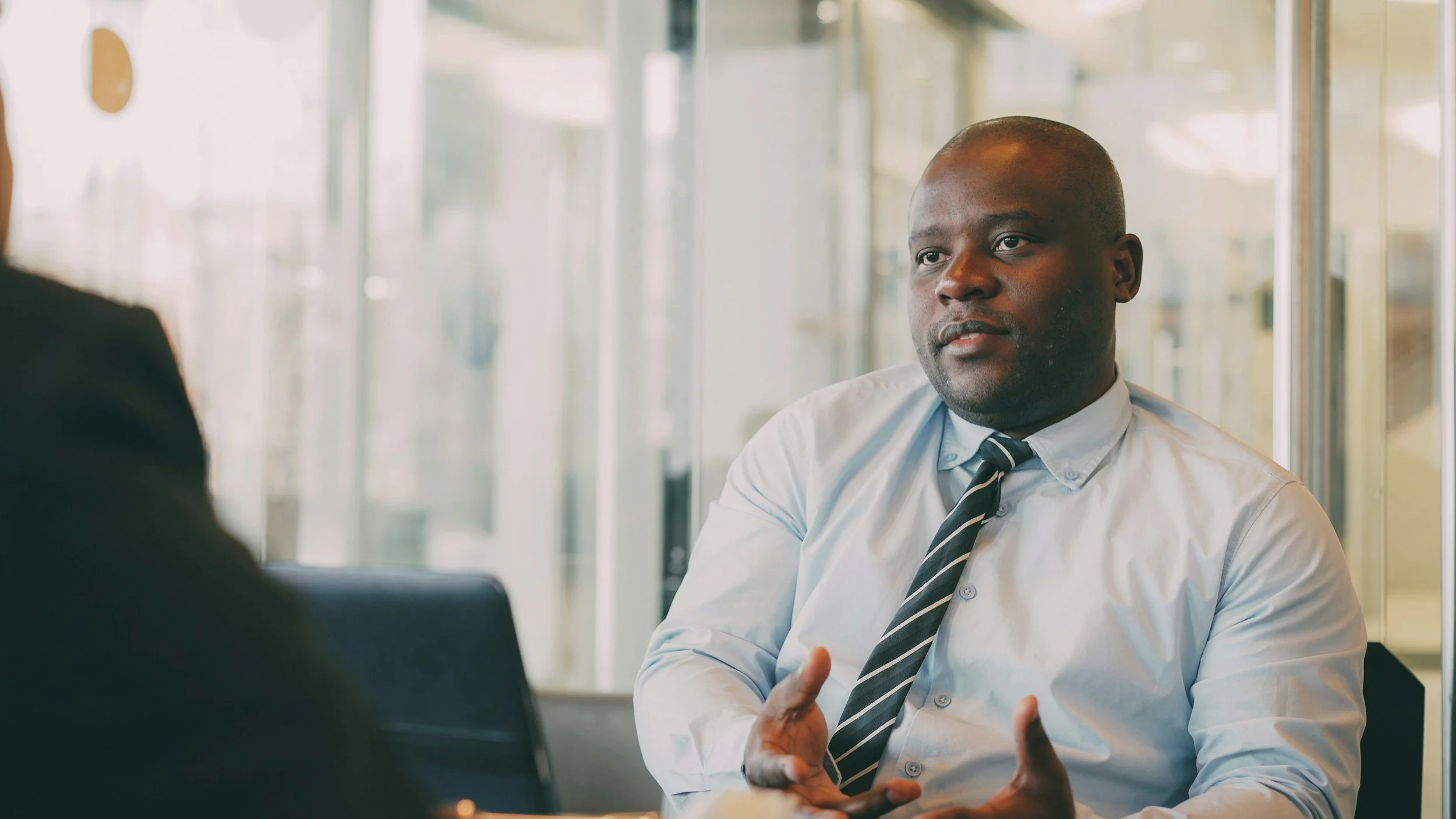 A man in a white shirt and striped tie is speaking and gesturing with his hands during a business meeting or interview in a modern office setting.