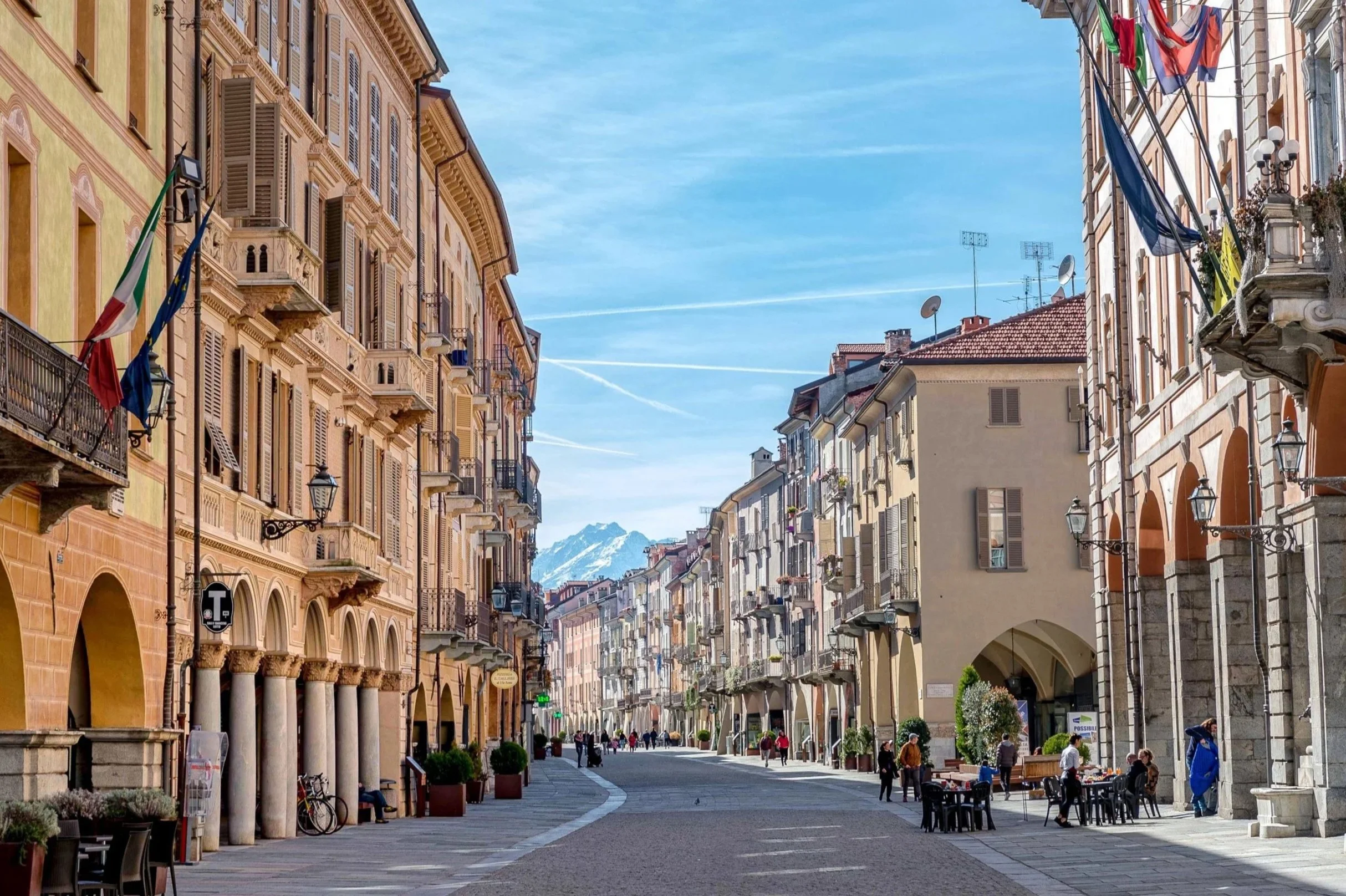 Una strada pedonale cittadina con edifici storici ai lati e alcune persone che passeggiano, con montagne innevate sullo sfondo, in una giornata soleggiata.